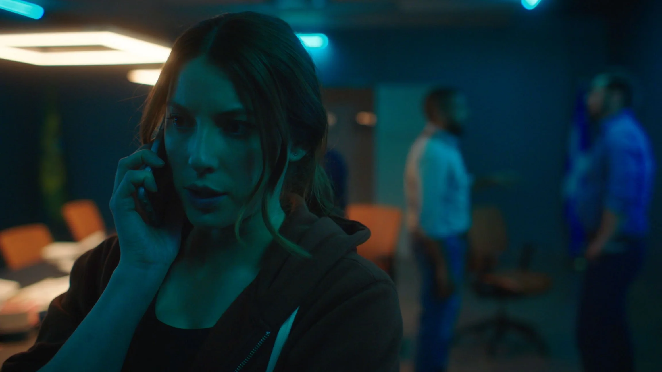 A woman with brown hair on the phone in a dimly lit office or conference room with blurred people in the background