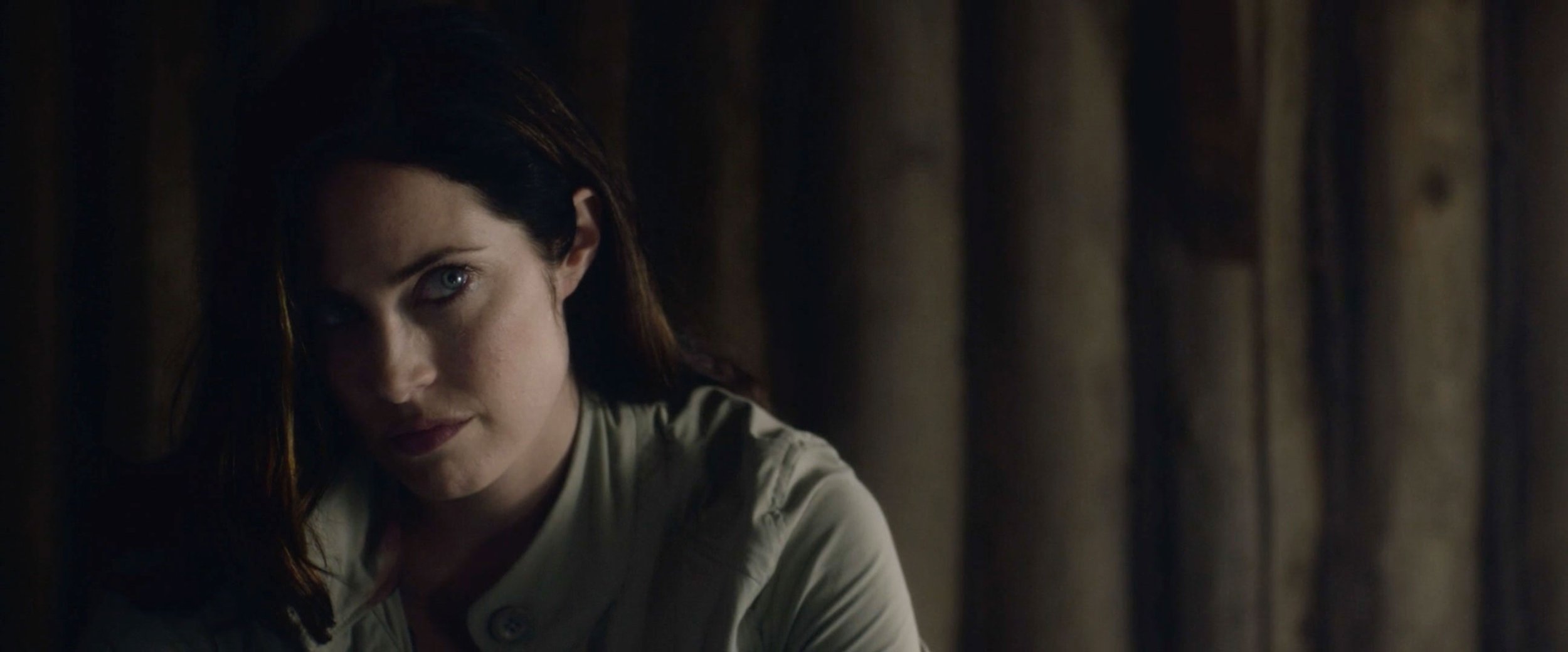 A close-up of a woman with dark hair and pale skin, looking at the camera with one eye visible, against a dark background with wooden panels.