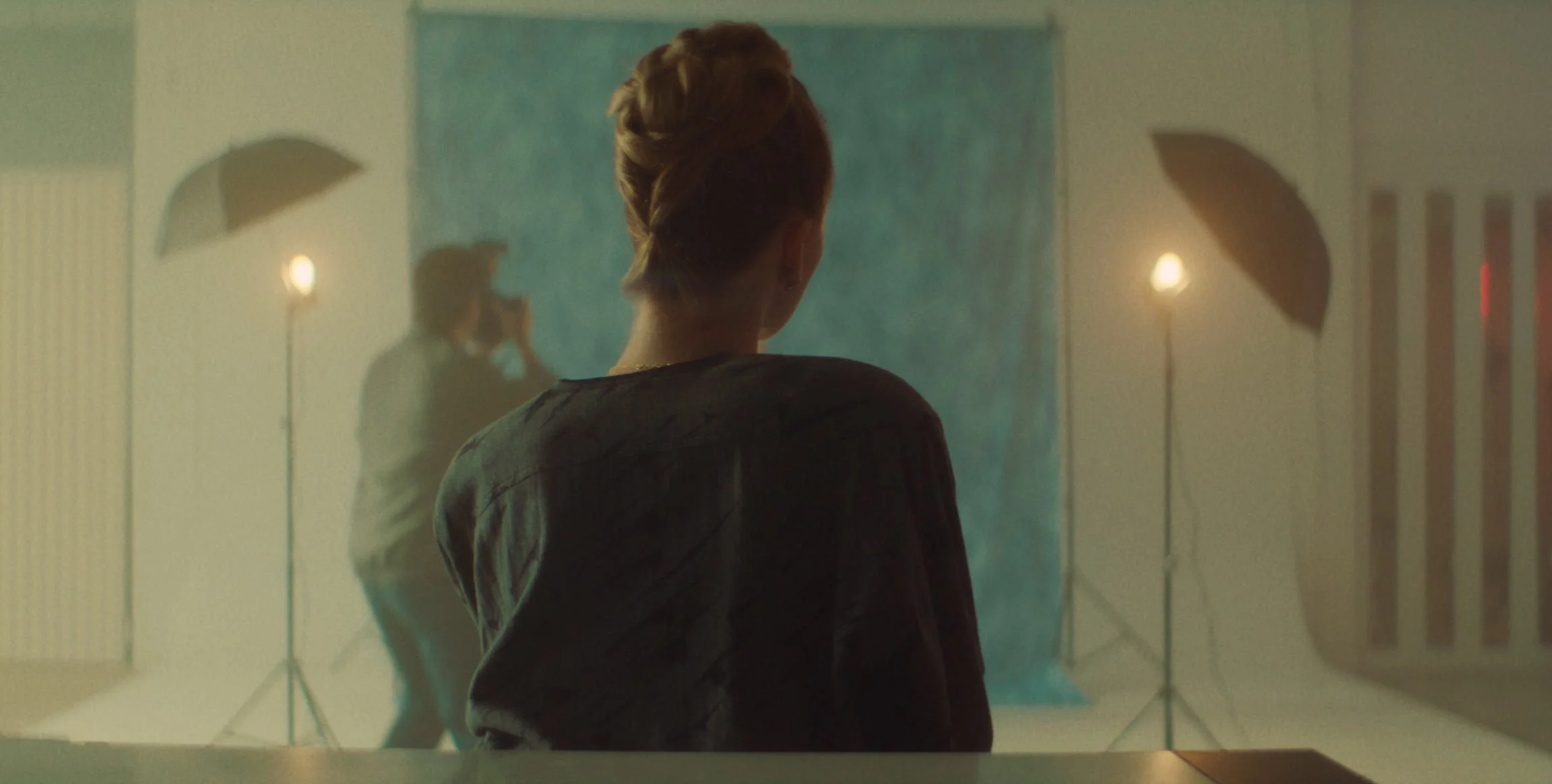 A woman with braided hair sitting and facing a photography studio setup with two studio lights, a blue backdrop, and another woman in the background talking on a phone.