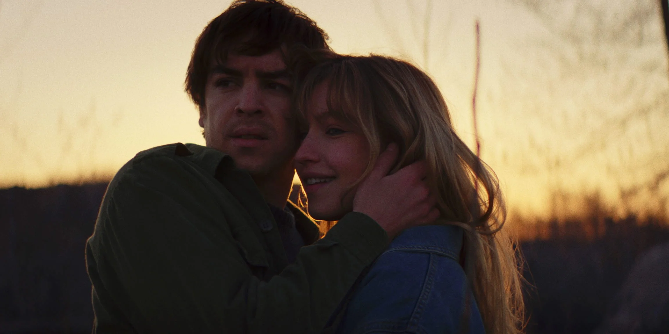 A man and woman embrace outdoors during sunset, looking emotional.