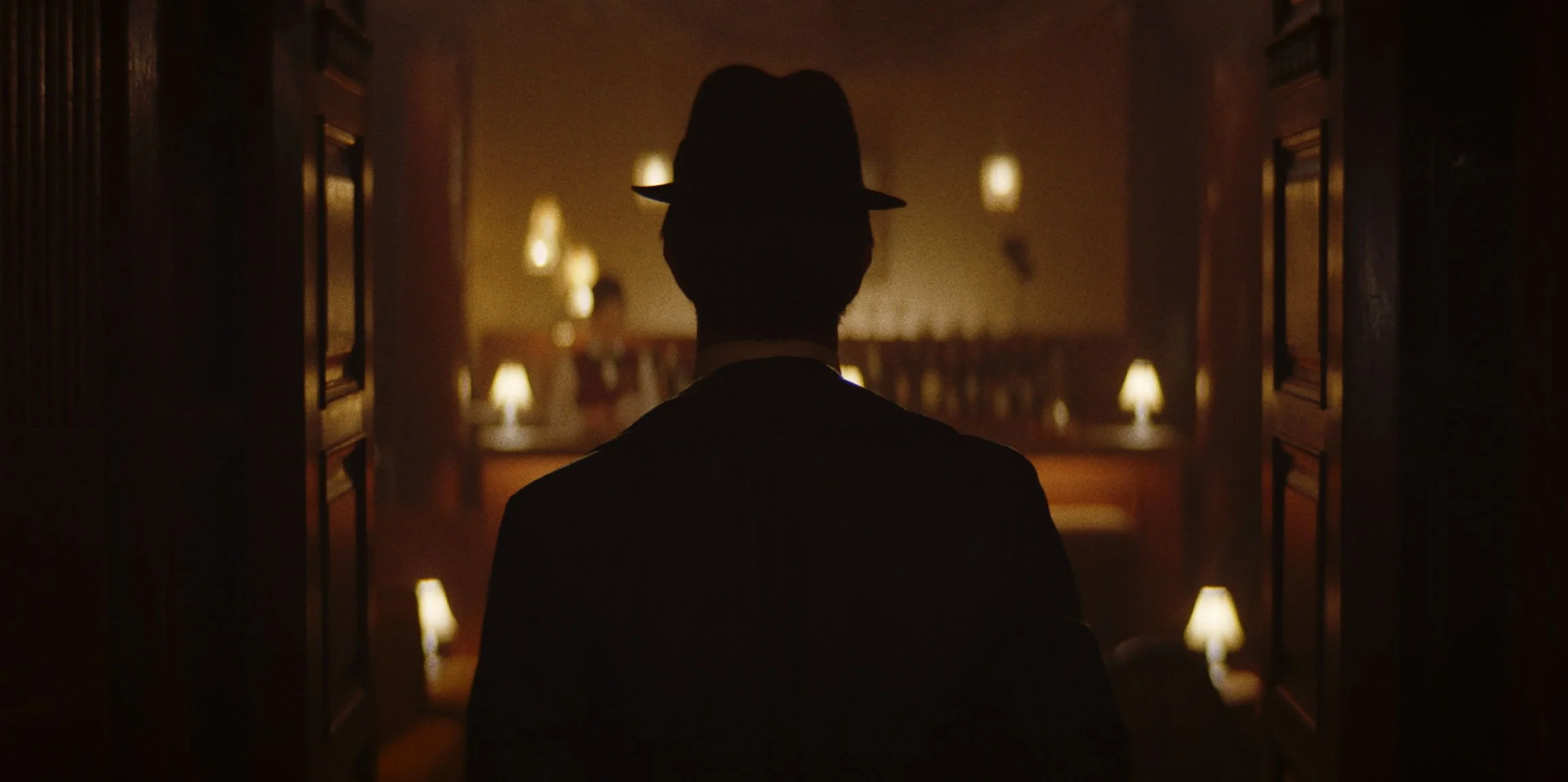 A silhouette of a man wearing a hat, sitting in a dimly lit room with lamps, viewed from behind as he faces a decorated wall with blurred figures in the background.