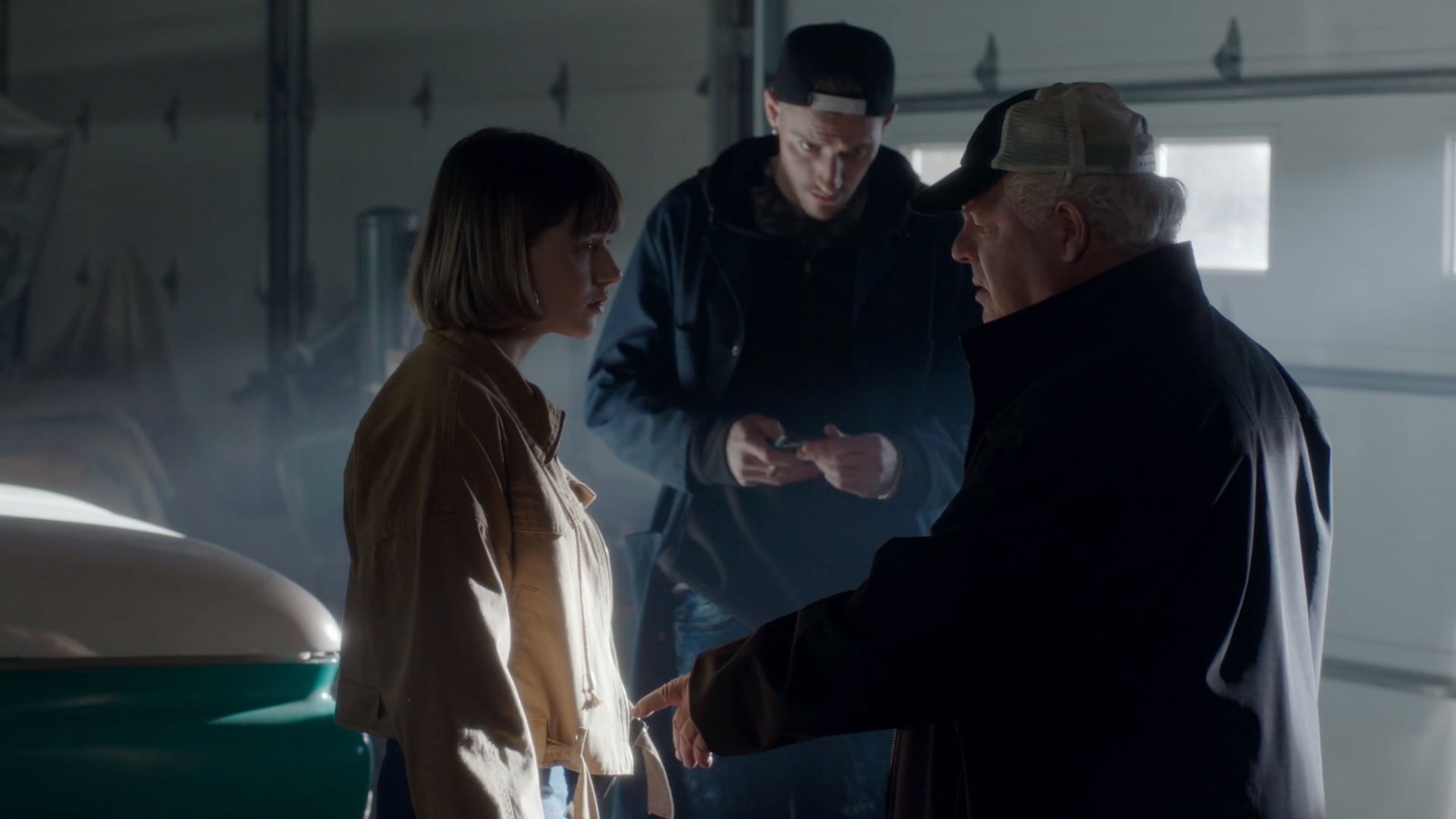 Three people in a garage with a vintage car, engaged in a serious conversation, with one person holding a phone and another making a gesture.