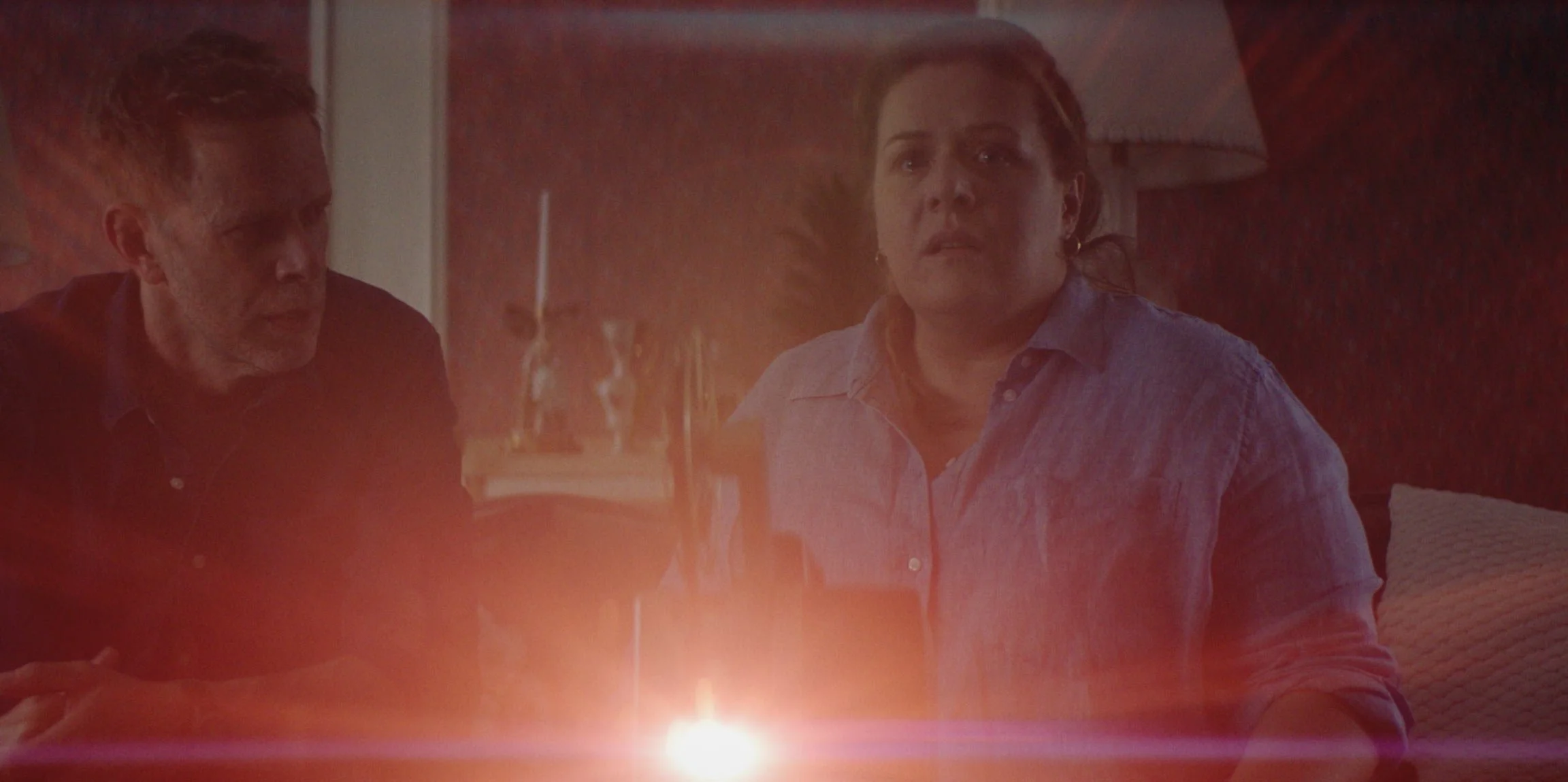 Two people sitting at a table illuminated by a candle, with a warm, cozy background and a lamp behind them.