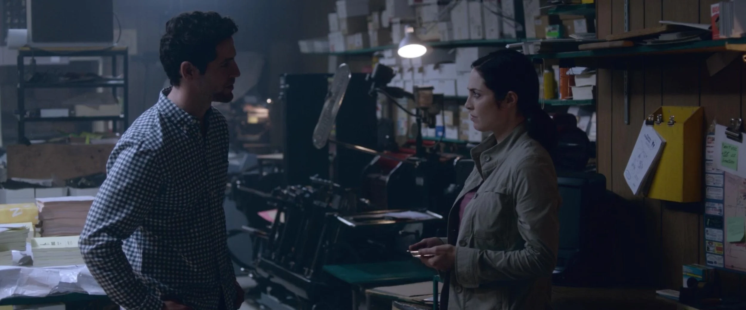 A man and woman are having a serious conversation in a cluttered office filled with papers, boxes, and office supplies.