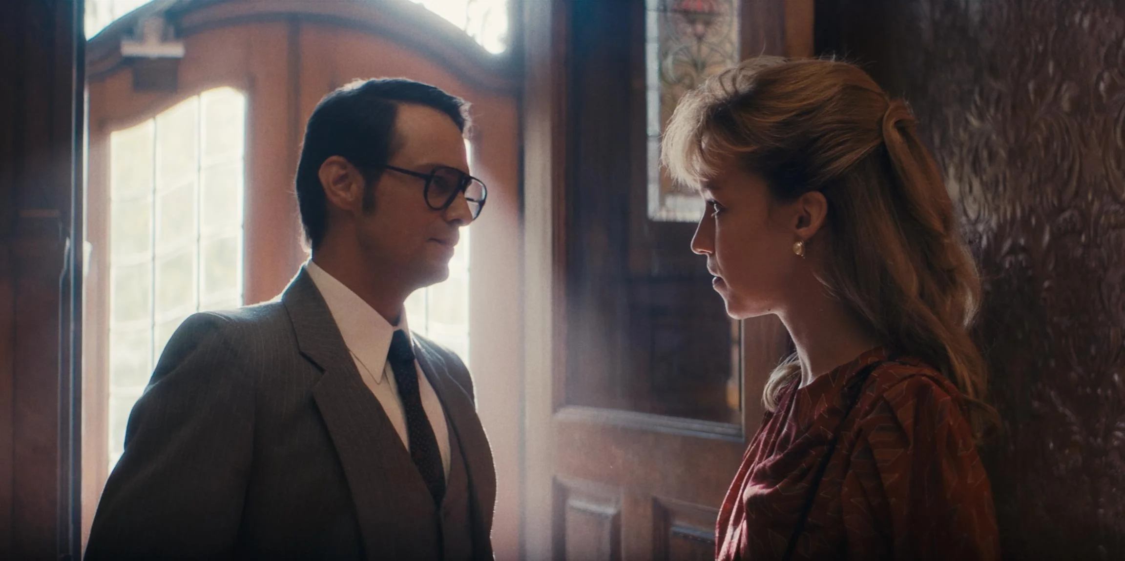 A man in a gray suit and glasses facing a woman with blonde hair inside a room with wooden walls and large window.