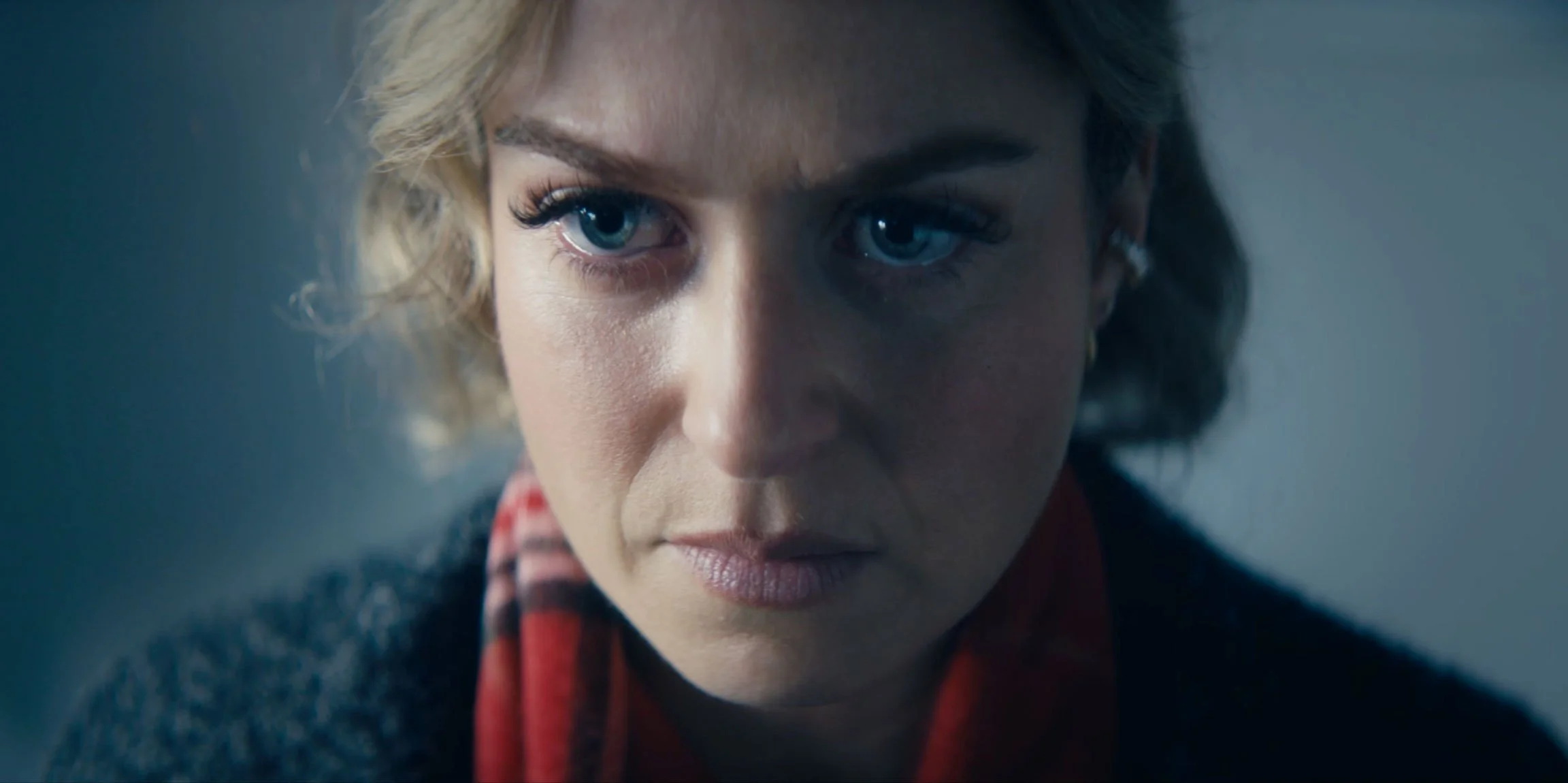 Close-up of a woman with blonde hair, blue eyes, and a serious expression, wearing a red scarf and a dark sweater.