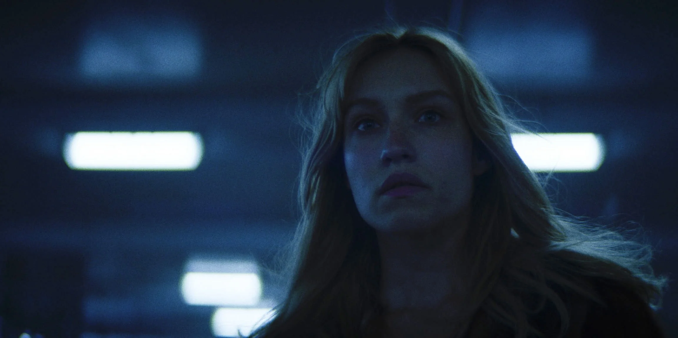A woman with long wavy hair stands in a dimly lit parking garage, looking forward with a serious expression.