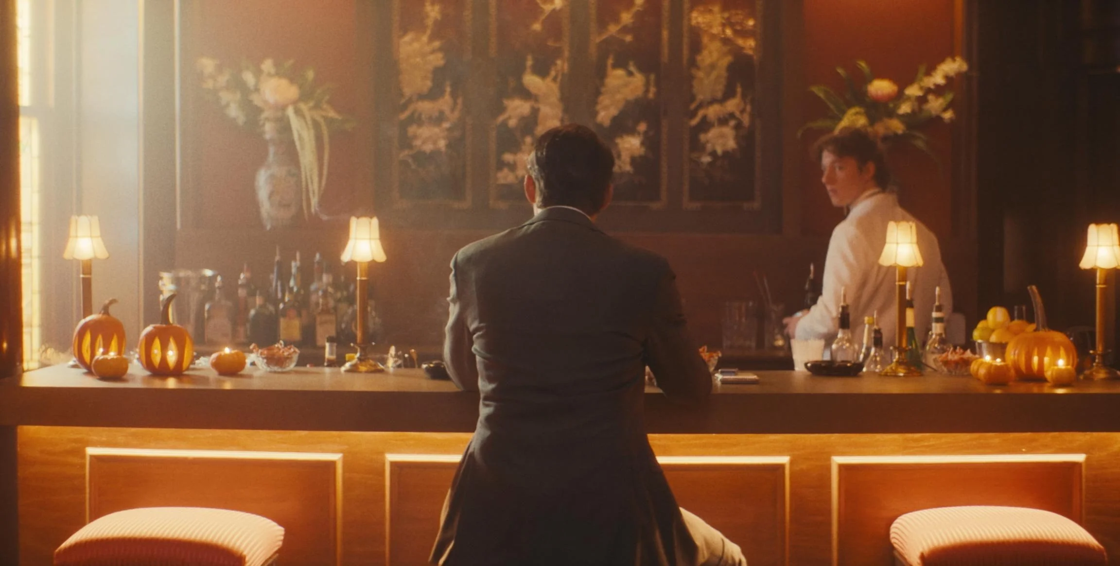 A man in a suit sitting at a bar with Halloween-themed decorations, pumpkins, and bar tools, while a woman wearing a white outfit stands behind the bar in a warmly lit, vintage-style setting.