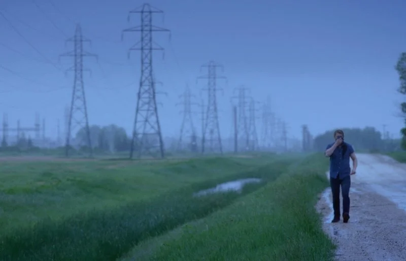 A person walking along a dirt path next to a grassy area with power lines and pylons in the background.