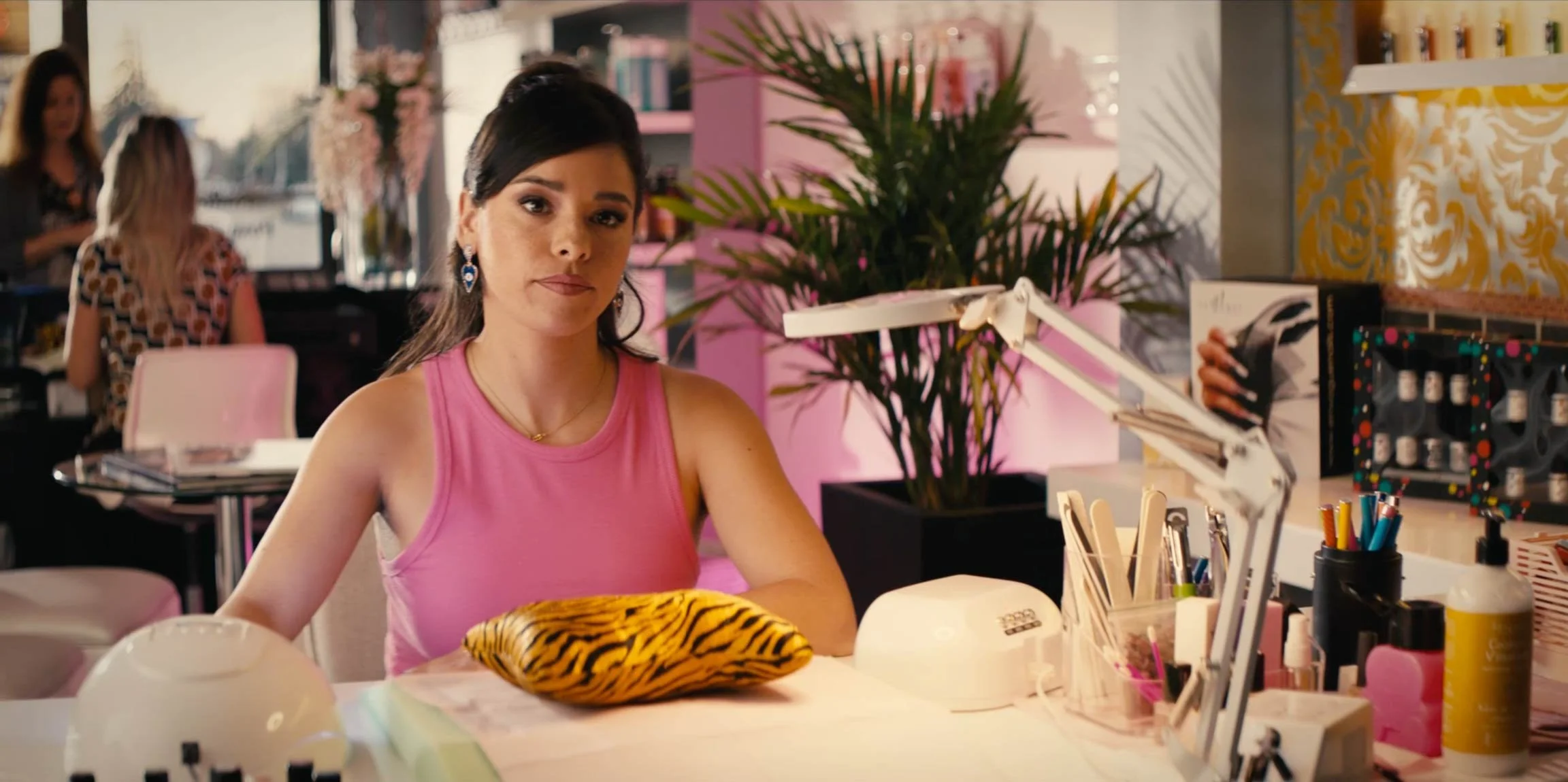 Woman in a pink sleeveless top sitting at a manicure station with various beauty tools, a tiger print cloth, and a lamp, inside a brightly decorated salon.