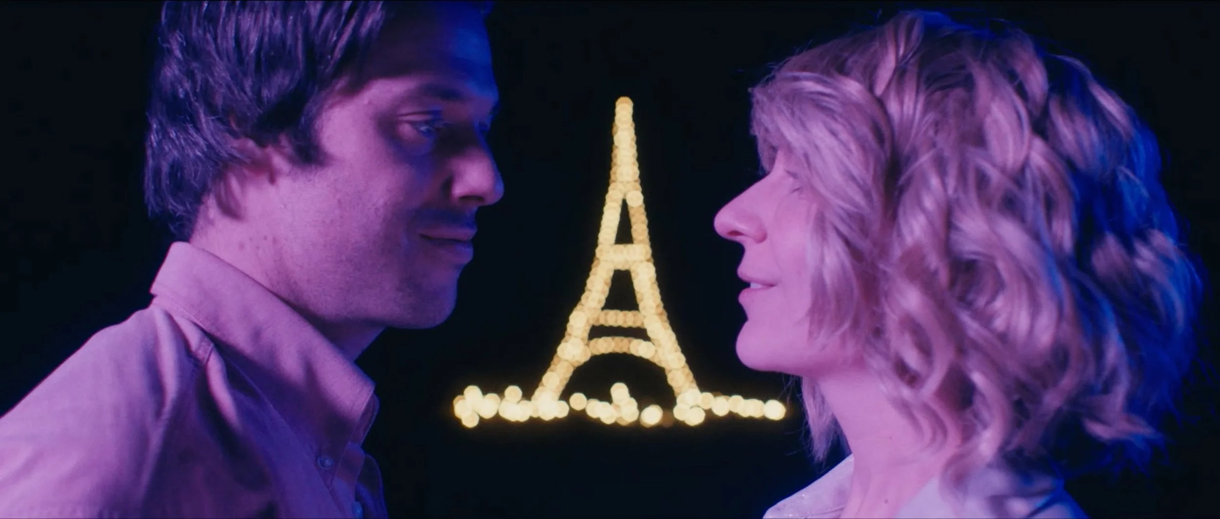 A young man and woman with their faces close together, facing each other, with the Eiffel Tower lit up in the background at night.