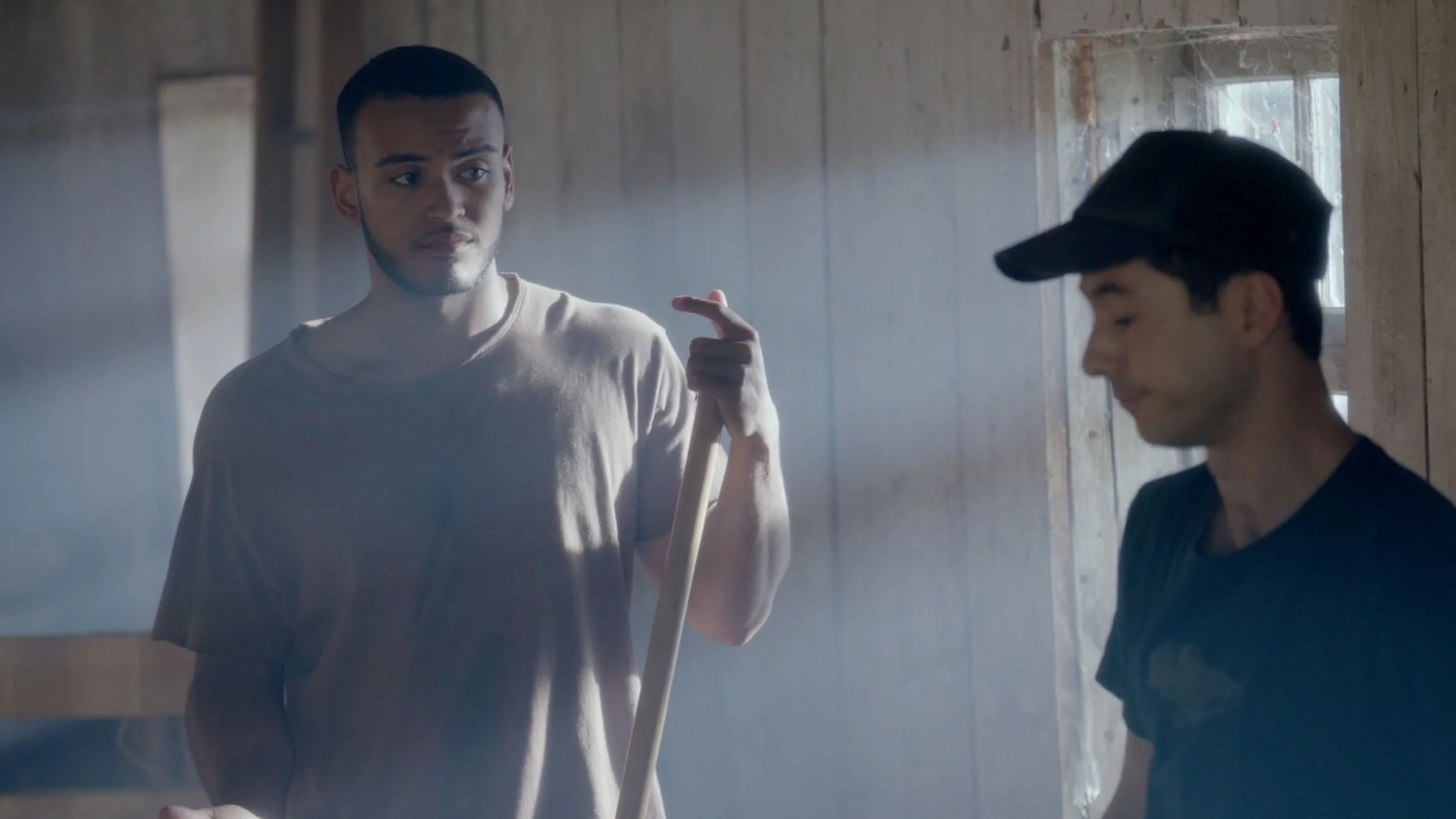 Two young men standing indoors, one holding a broom handle and pointing as if giving instructions, both appearing to be in conversation, with natural light coming through a window.