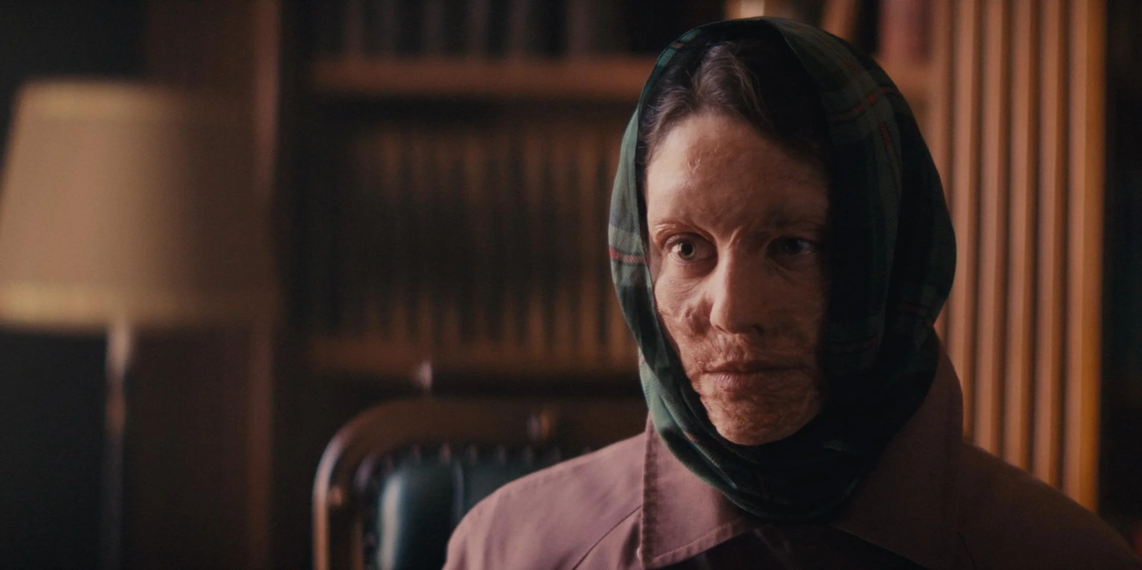 A woman wearing a headscarf with a plaid pattern, sitting in a dimly lit room with wooden paneling, looking to the side with a serious expression.