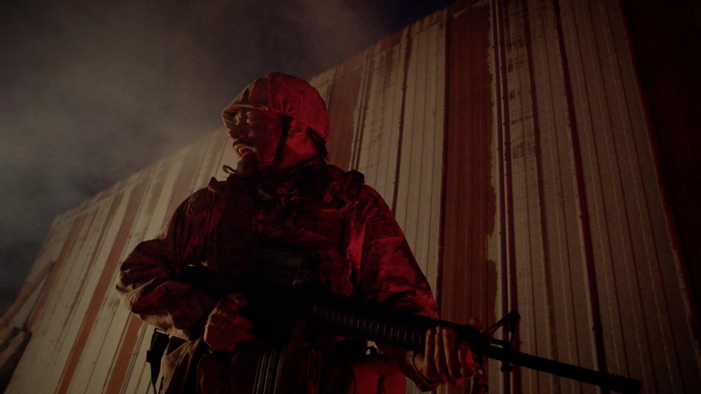 A person dressed in a red camouflage jacket and hood, holding a rifle, standing against a dark, cloudy sky and wooden wall.