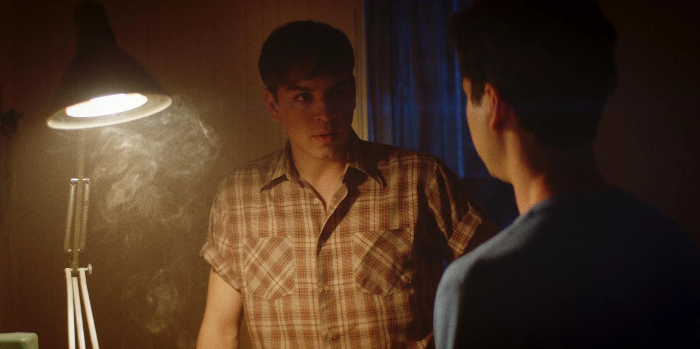 Two young men having a serious conversation in a dimly lit room, with a lamp nearby and visible smoke in the air.