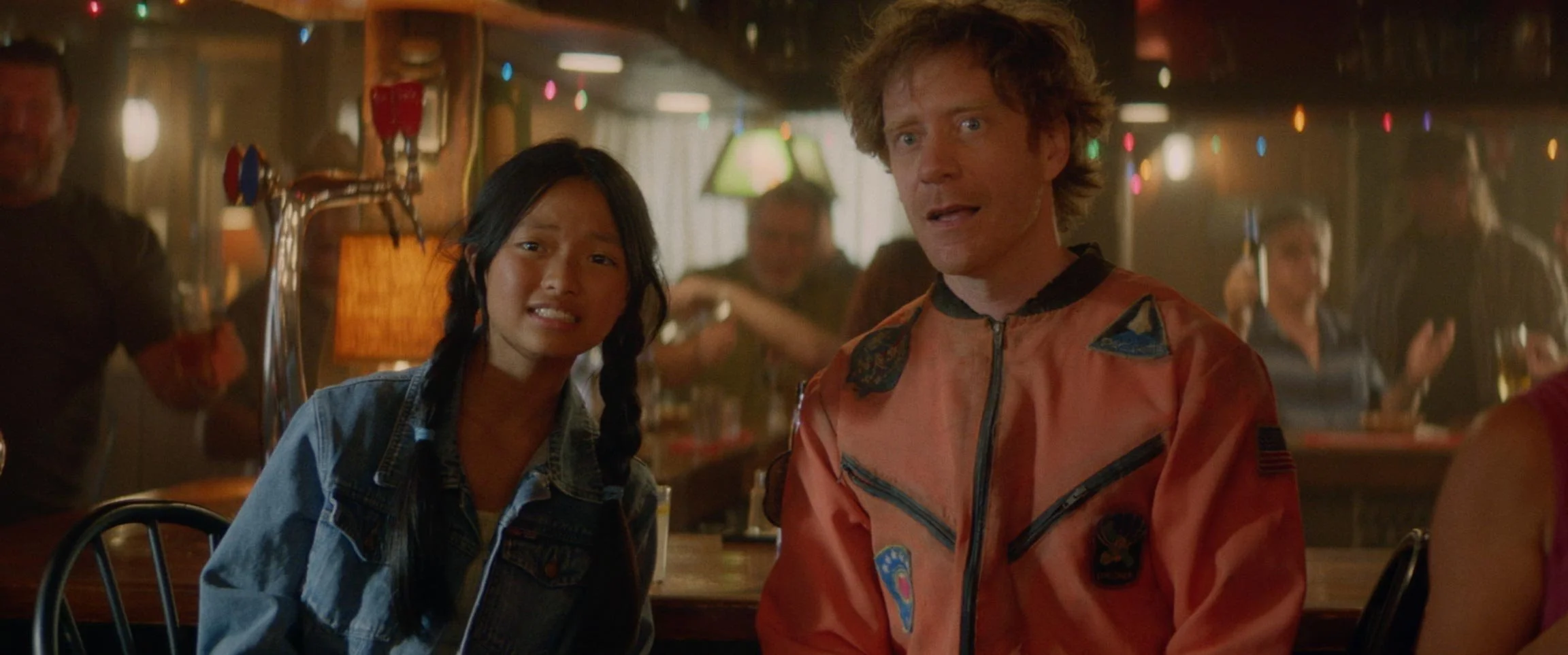 A woman and a man sitting at a bar, looking surprised or confused, with colorful lights in the background.