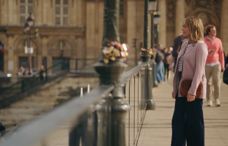 A woman in a pink jacket and striped shirt standing on a city sidewalk, looking toward the water with a historic building in the background.