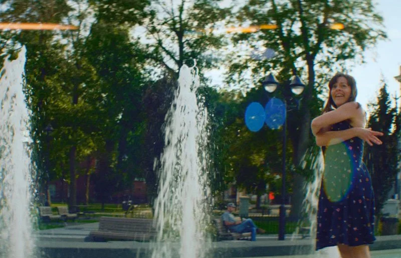 A woman smiling and hugging herself near a fountain in a park with trees and benches.