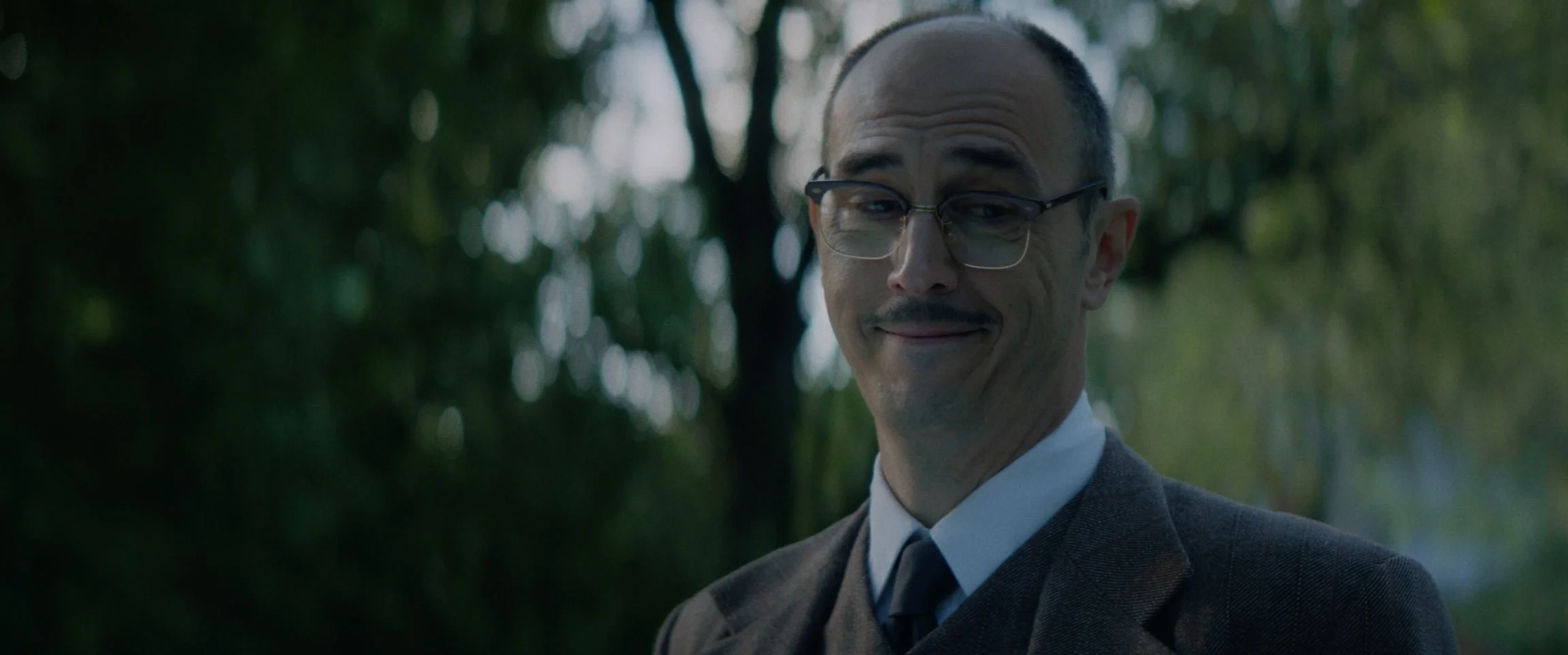 A man wearing glasses, a suit, and a tie, smiling outdoors with trees in the background.
