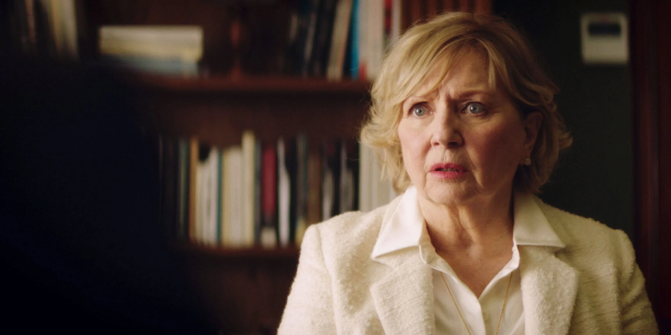 A middle-aged woman with blonde hair, wearing a white blouse and blazer, appears surprised or concerned as she looks to the side in an indoor setting with a bookshelf in the background.