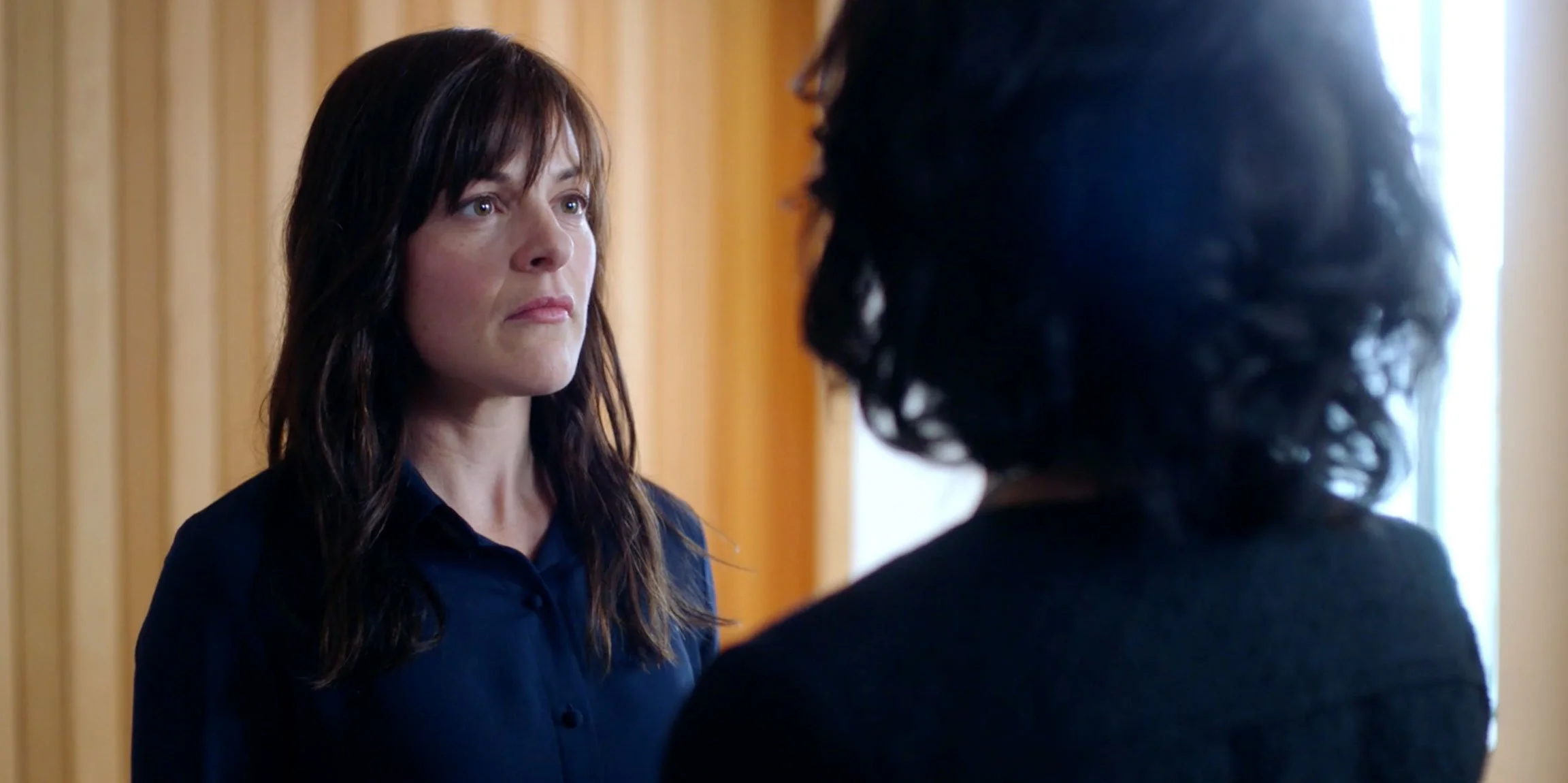 A woman with dark brown hair and a serious expression facing another woman with curly black hair, in a room with wooden paneling and bright natural light.
