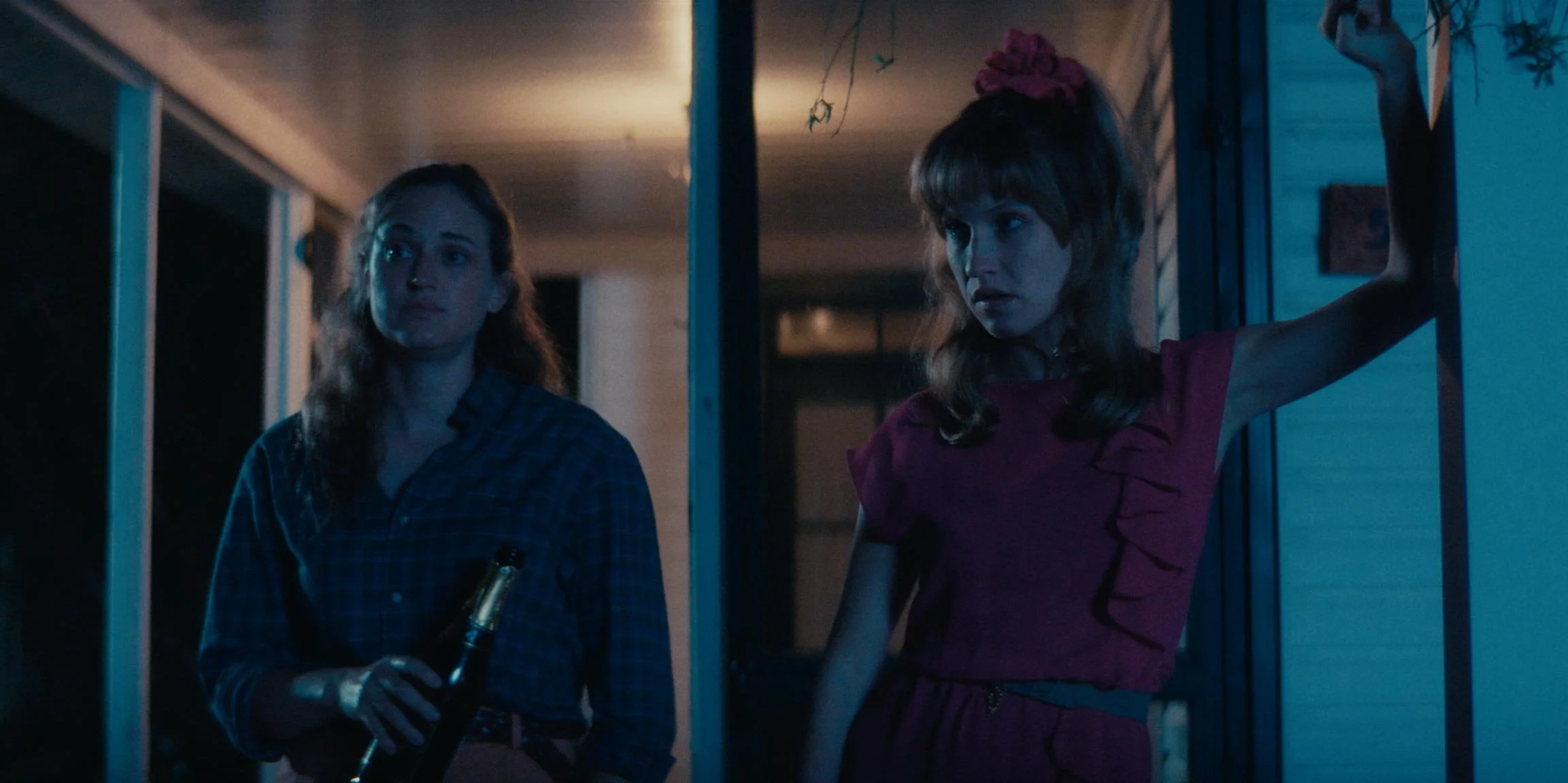 Two women standing on a porch at night, one holding a bottle, and the other leaning against the wall with her arm raised.