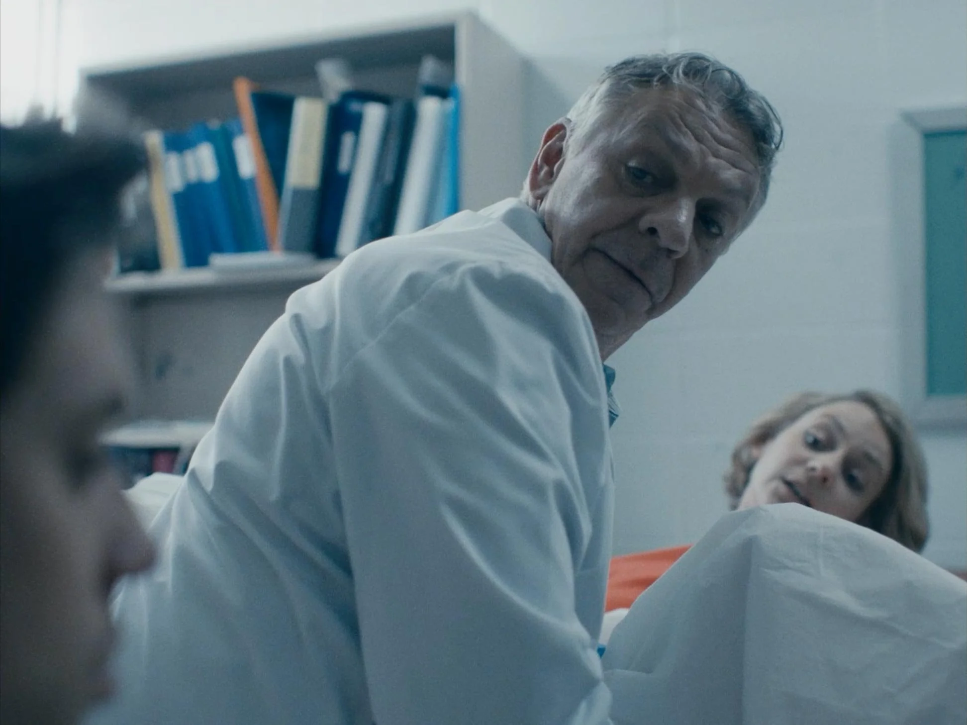 An older man in a white coat looks over his shoulder towards a woman and a young person, sitting on a bed in a hospital room with a bookshelf in the background.