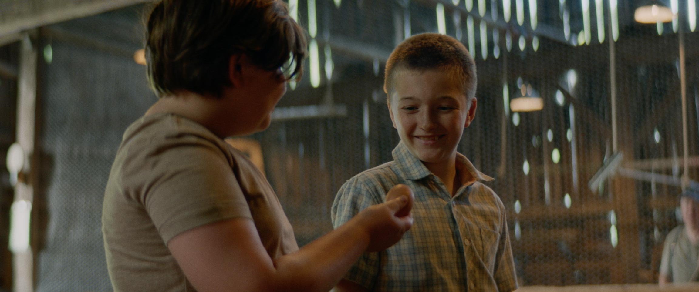 A young boy smiling while holding a baseball in a rustic indoor setting with two other children partially visible.