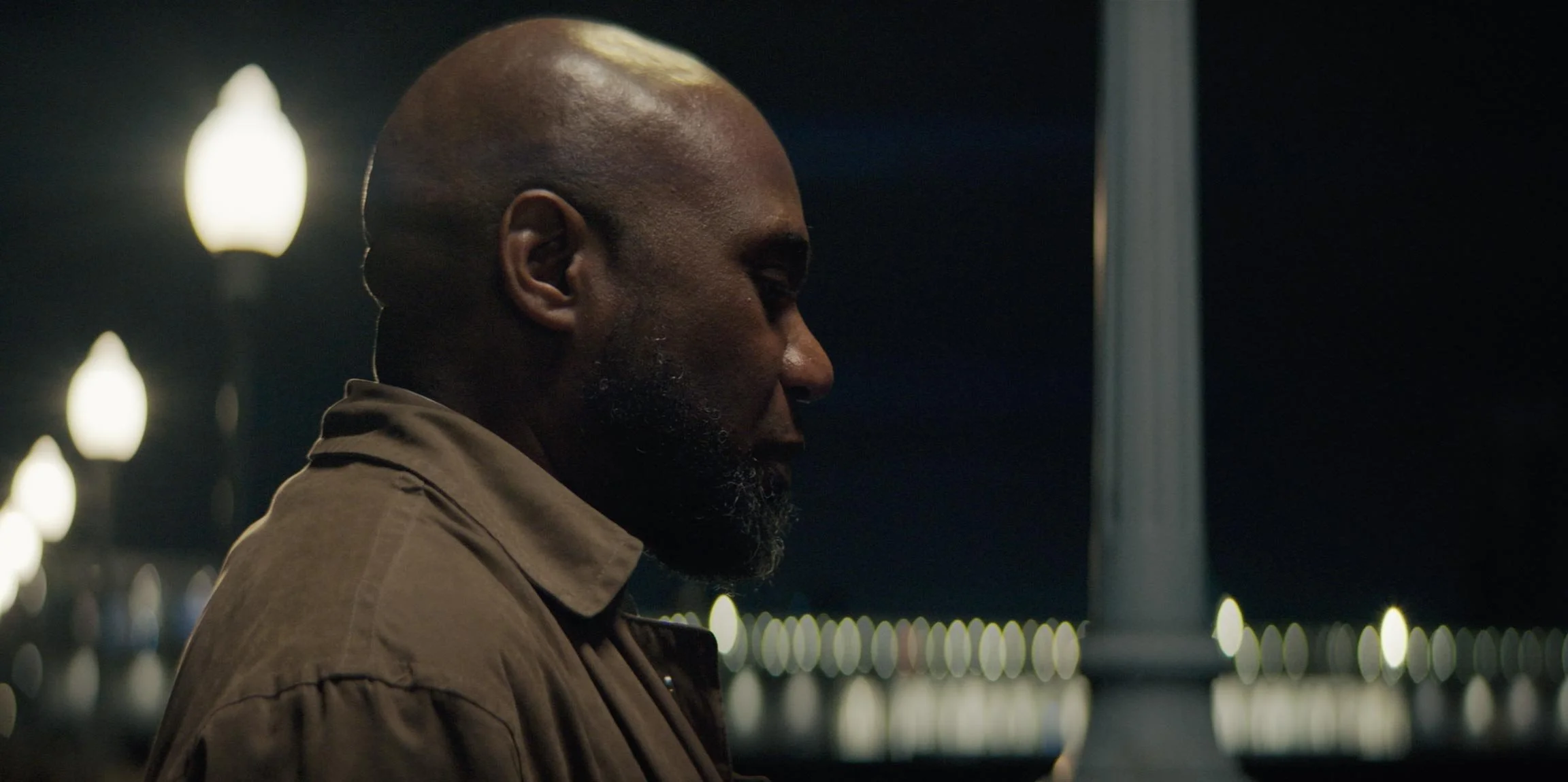 Profile of a man with a beard, standing outdoors at night, illuminated by streetlights, next to a lamp post.