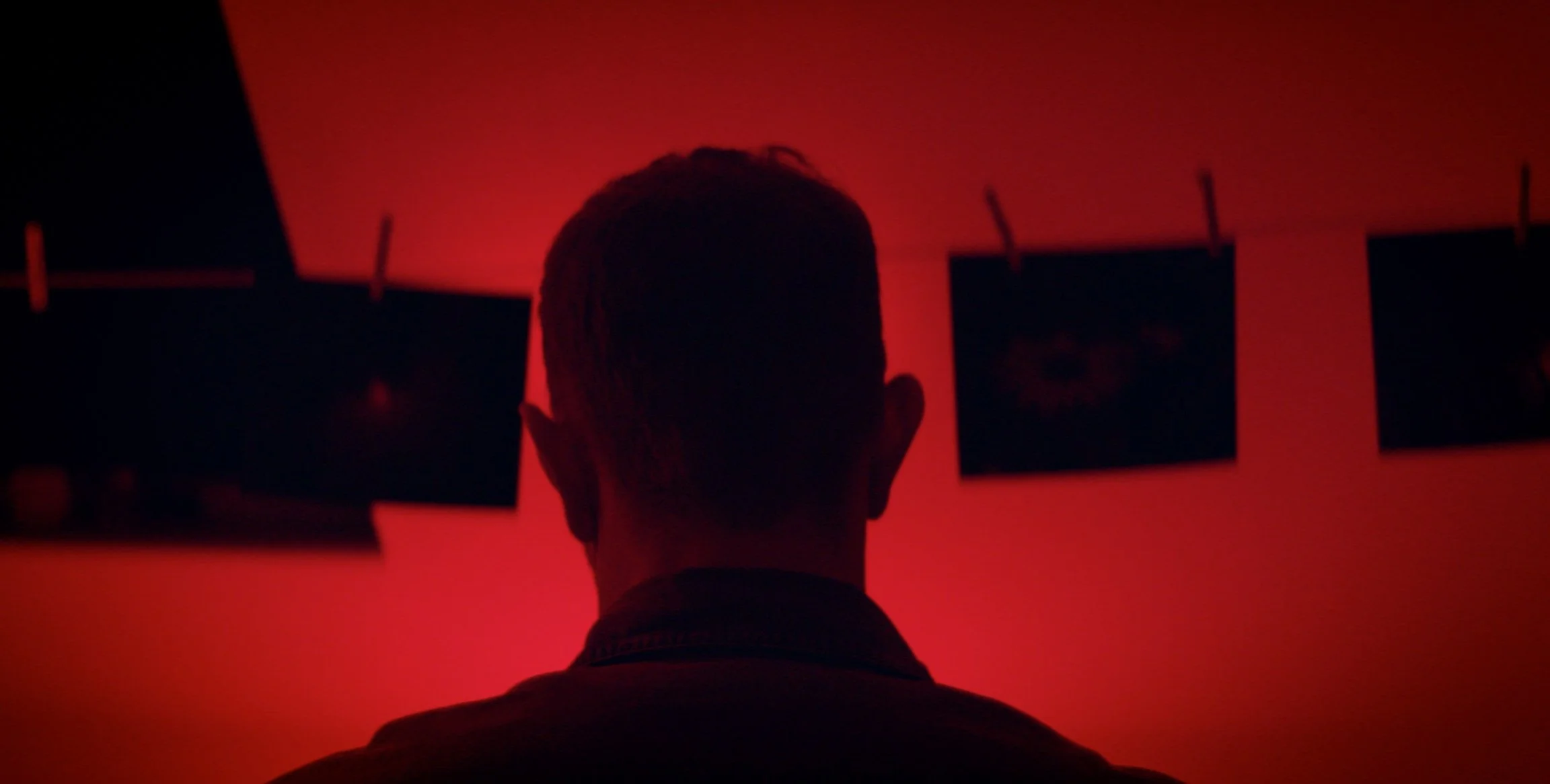 Back of a person's head in silhouette, facing a wall with three black-framed pictures hanging, illuminated by red light.