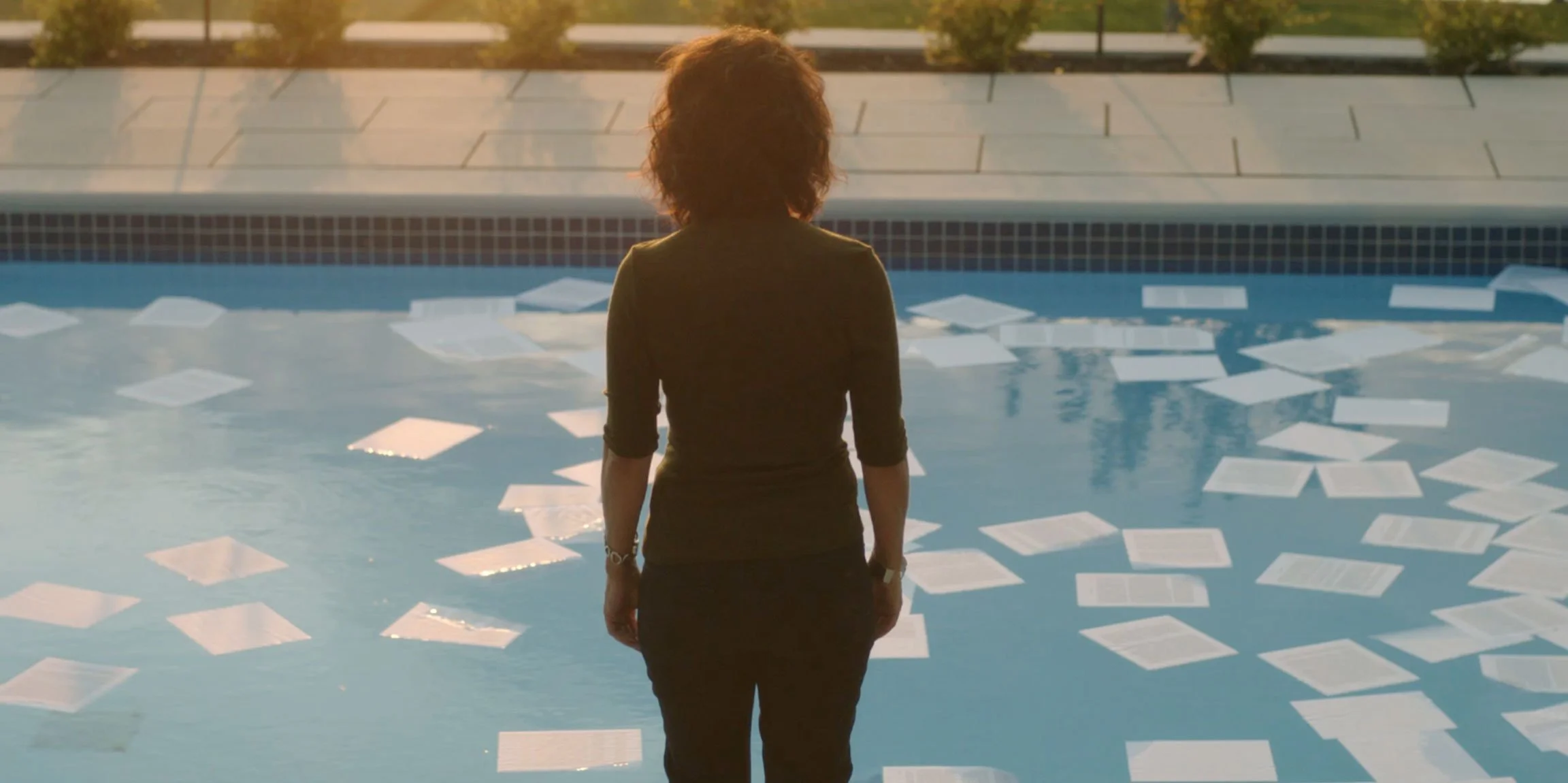 A woman standing by a swimming pool, looking at scattered papers floating on the water, during sunset.