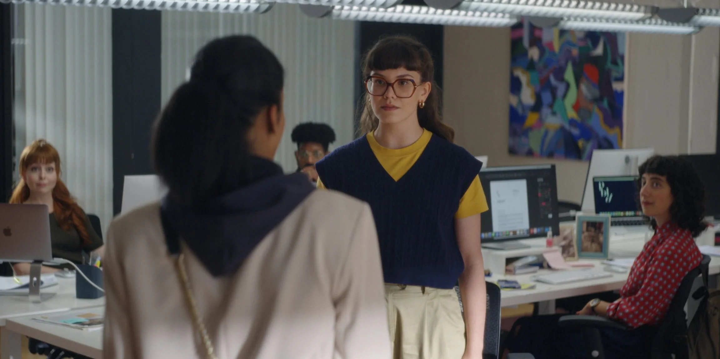 A woman with glasses and brown hair pulled back, wearing a yellow T-shirt and blue vest, stands in front of two women sitting at desks in an office. Other coworkers are visible in the background, working at computers.