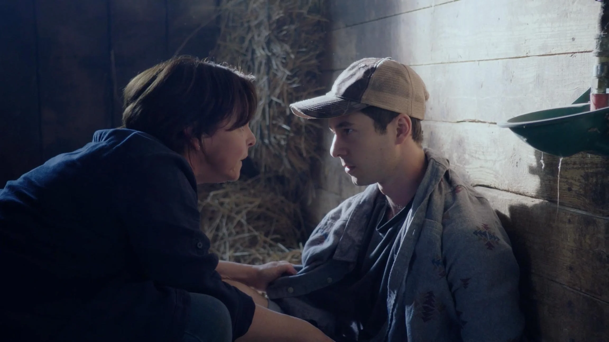 A woman and a young man are having a serious conversation indoors, sitting against a wooden wall with straw in the background.