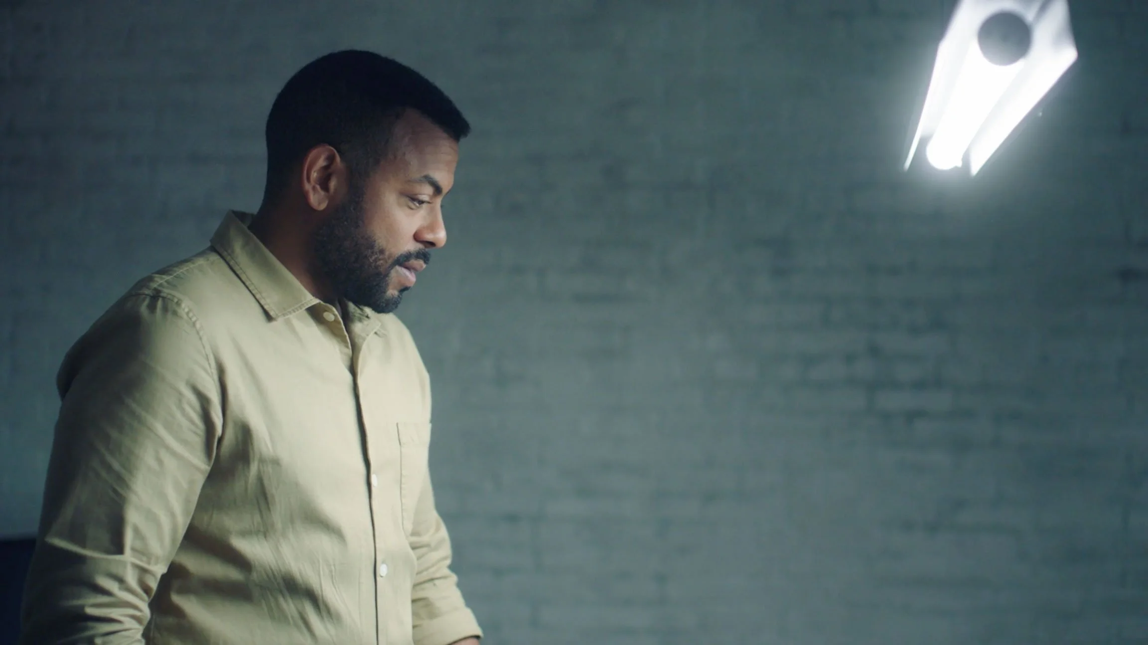 A man with a beard wearing a beige button-up shirt, looking down, in a room with a gray brick wall and fluorescent lighting.