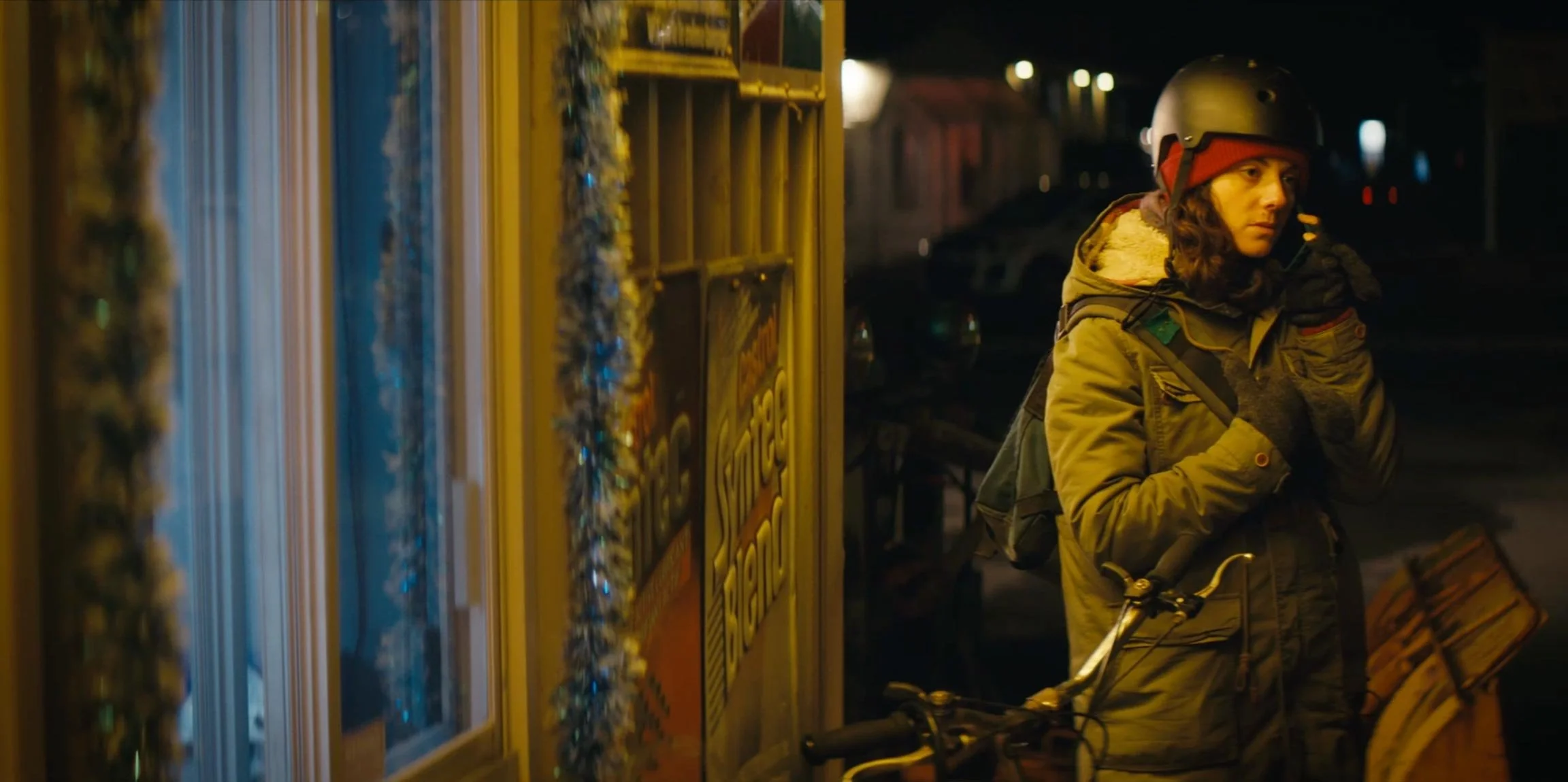 A woman with curly hair wearing a helmet and a green winter jacket stands next to a yellow phone booth decorated with tinsel, holding the handlebar of a bicycle at night.