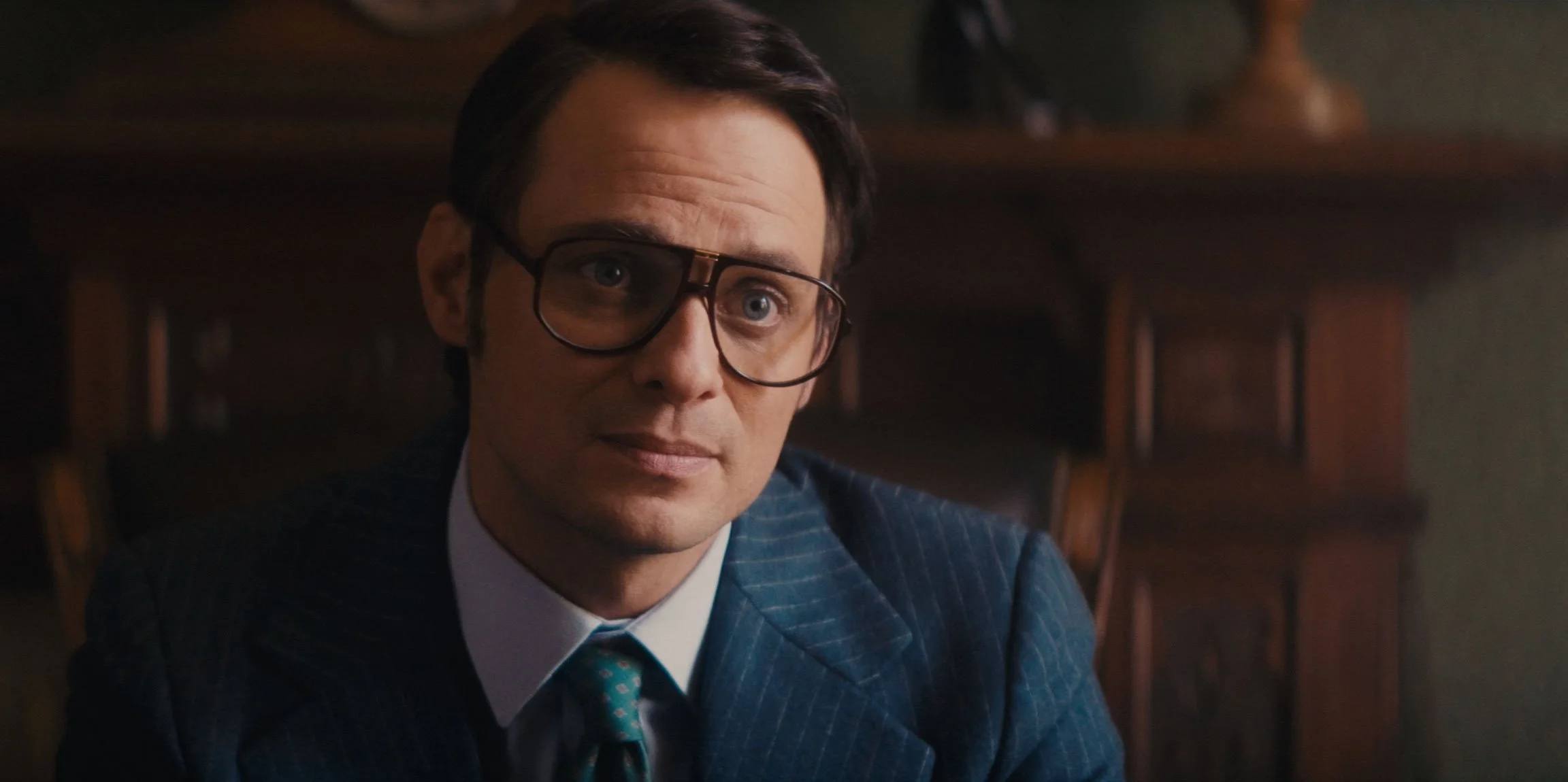 A man with dark hair, glasses, and wearing a pinstripe suit sitting at a desk in a room with wooden furniture.