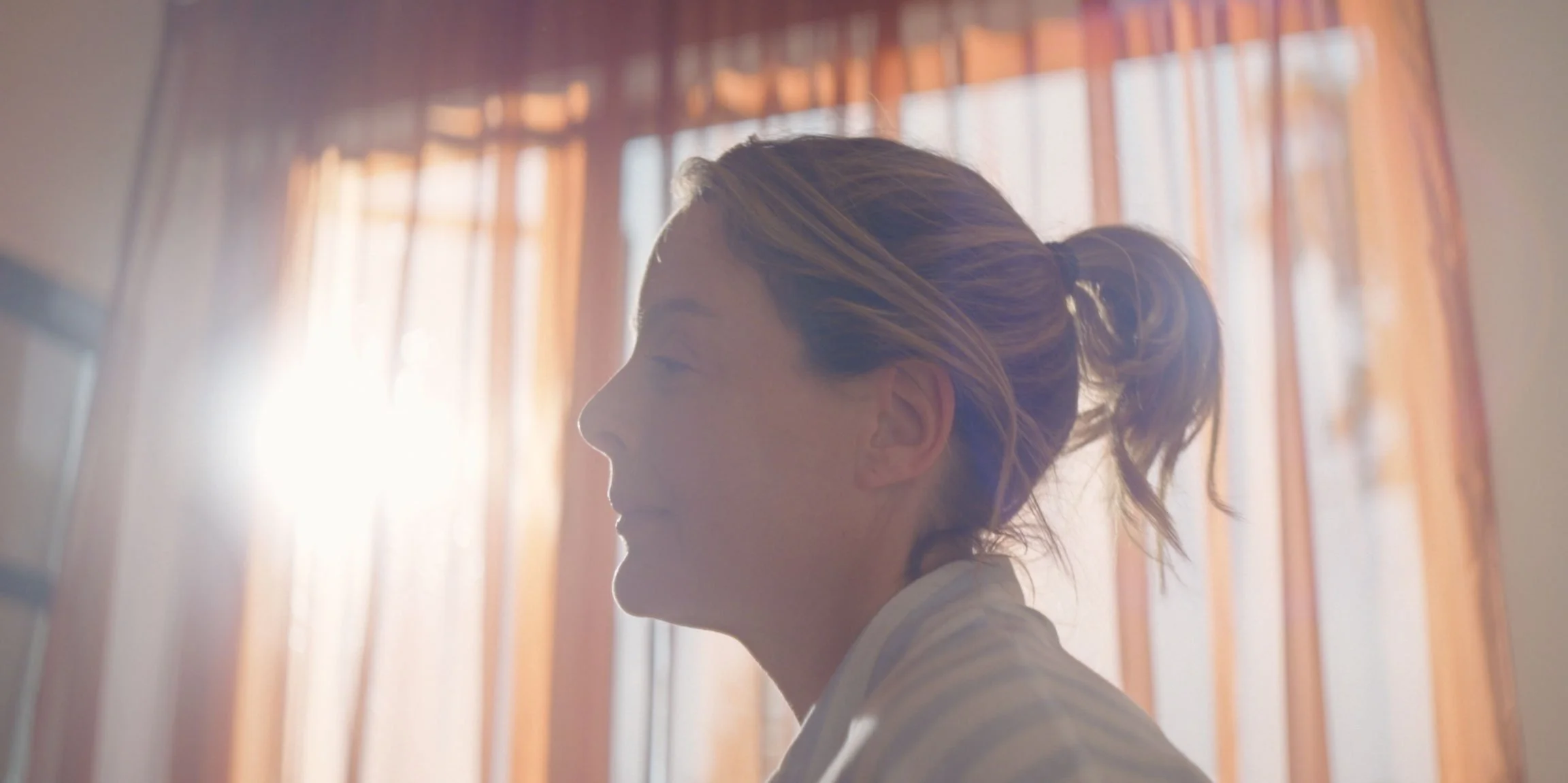 Side profile of a woman with blonde hair in a ponytail, standing in front of orange curtains, backlit by sunlight