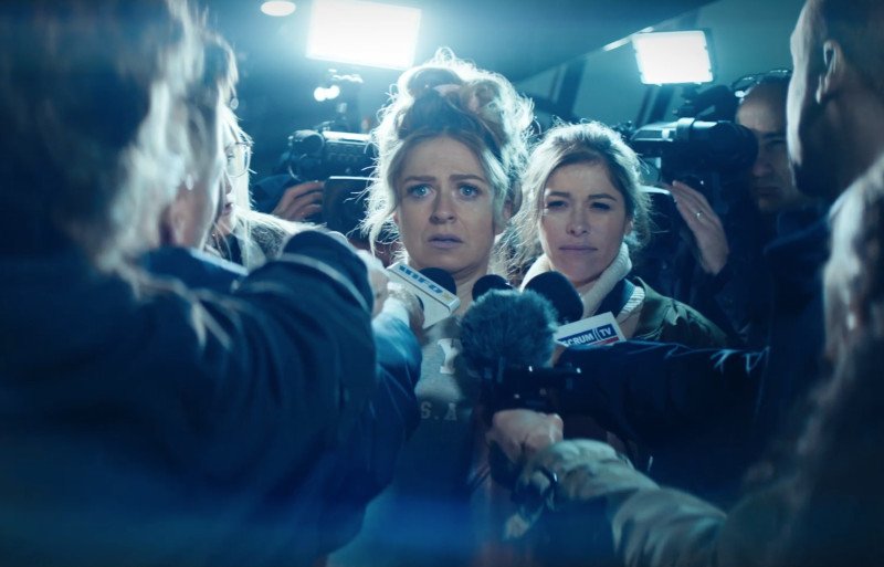 A woman surrounded by reporters and cameras at a press conference or interview in a crowded indoor setting.