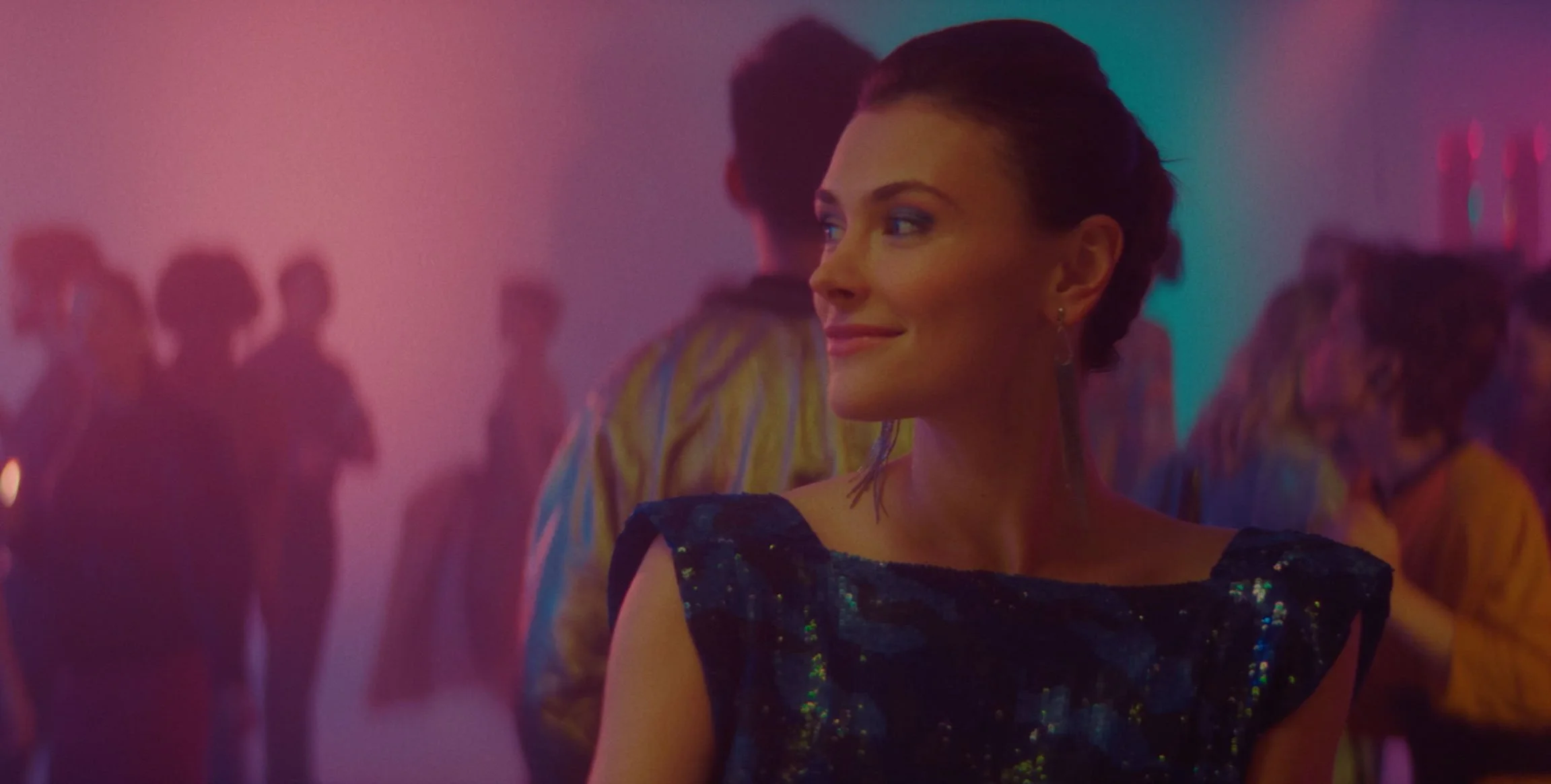 A woman with short dark hair, wearing a dark, sequined dress, smiling and looking to her right, in a colorful party or nightclub scene with blurred figures in the background.