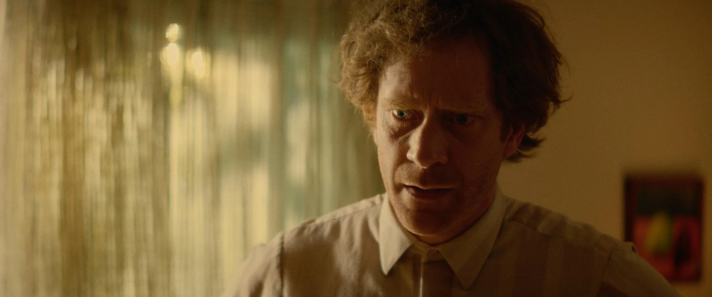 Close-up of a man with curly hair and a surprised or concerned expression, wearing a beige shirt, in a room with green curtains and warm lighting.