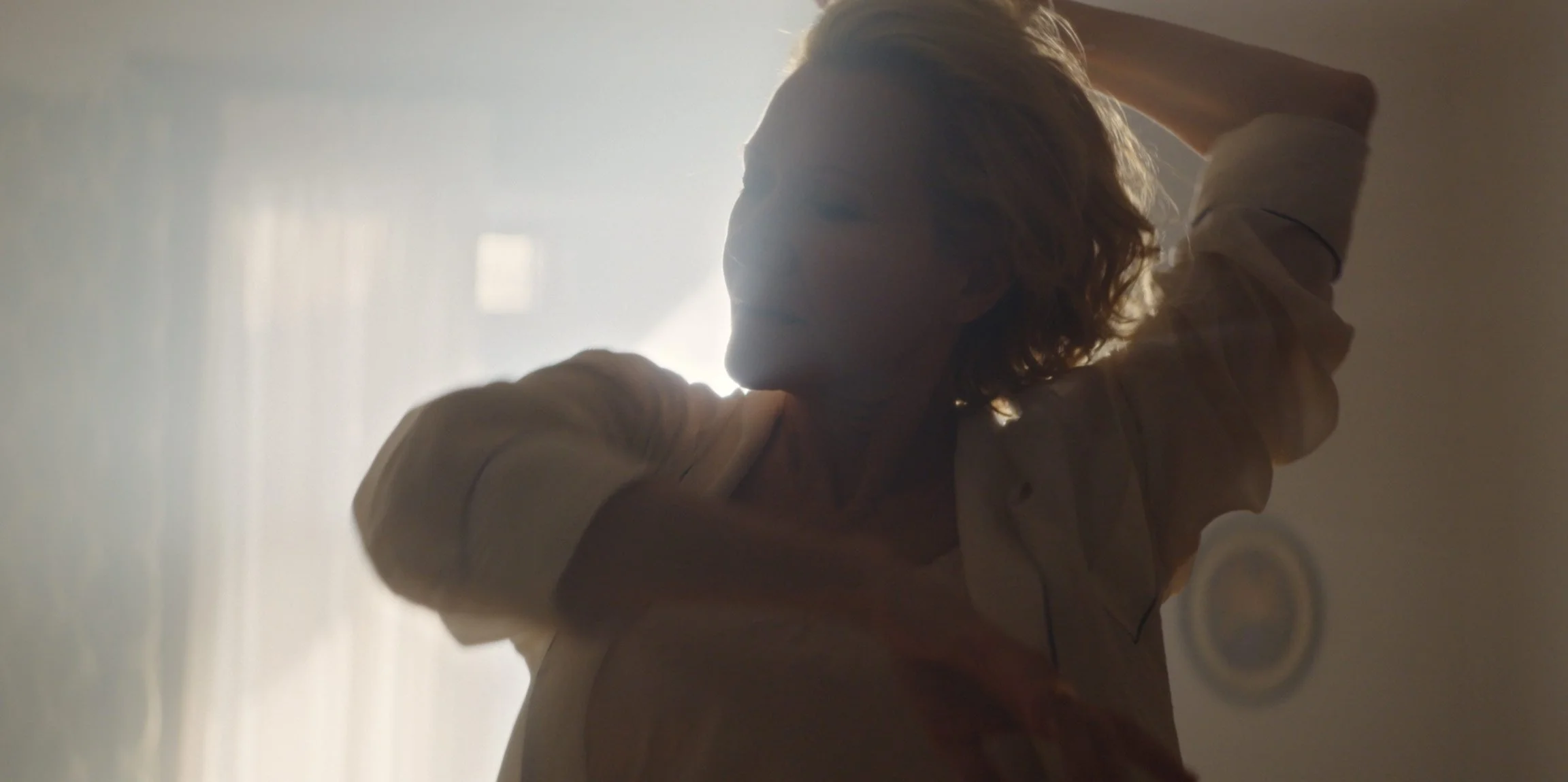A woman with short hair smiling and raising her arms, backlit by soft natural light.