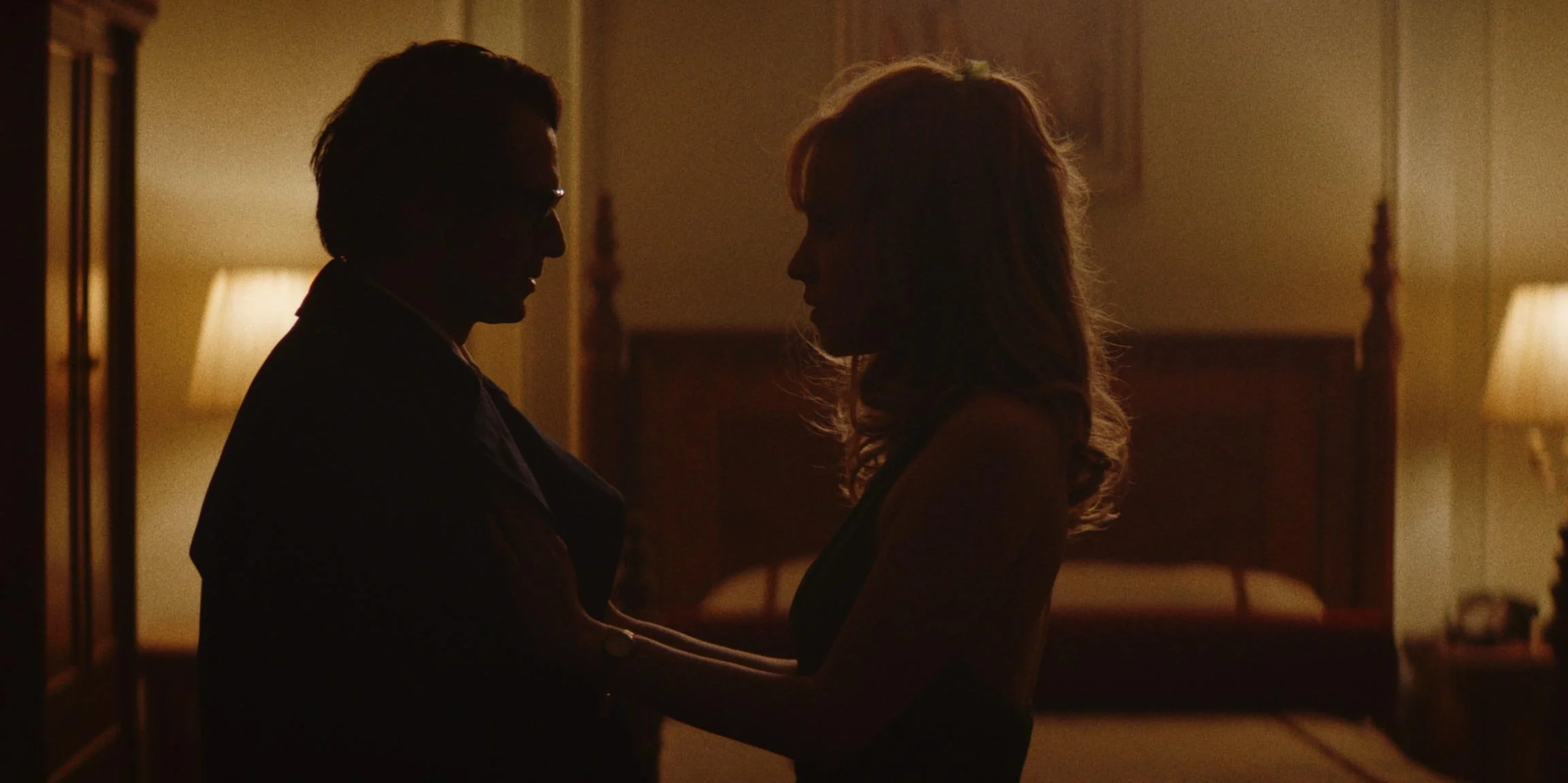 Silhouetted man and woman facing each other in a dimly lit bedroom, holding hands gently.