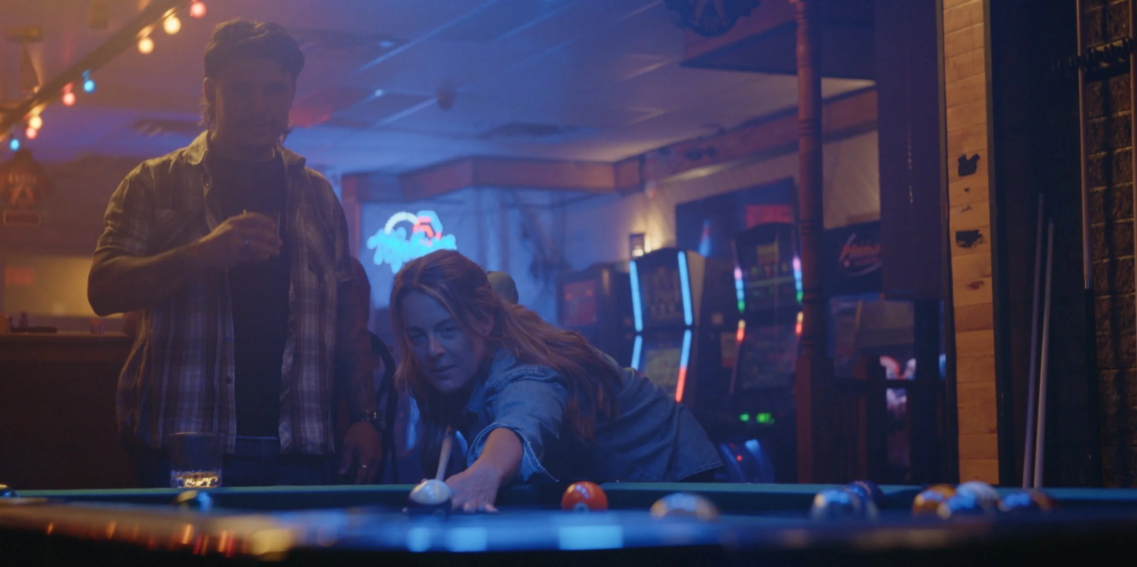 A woman plays pool at a bar, while a man stands nearby holding a drink, with colorful neon signs and arcade machines in the background.