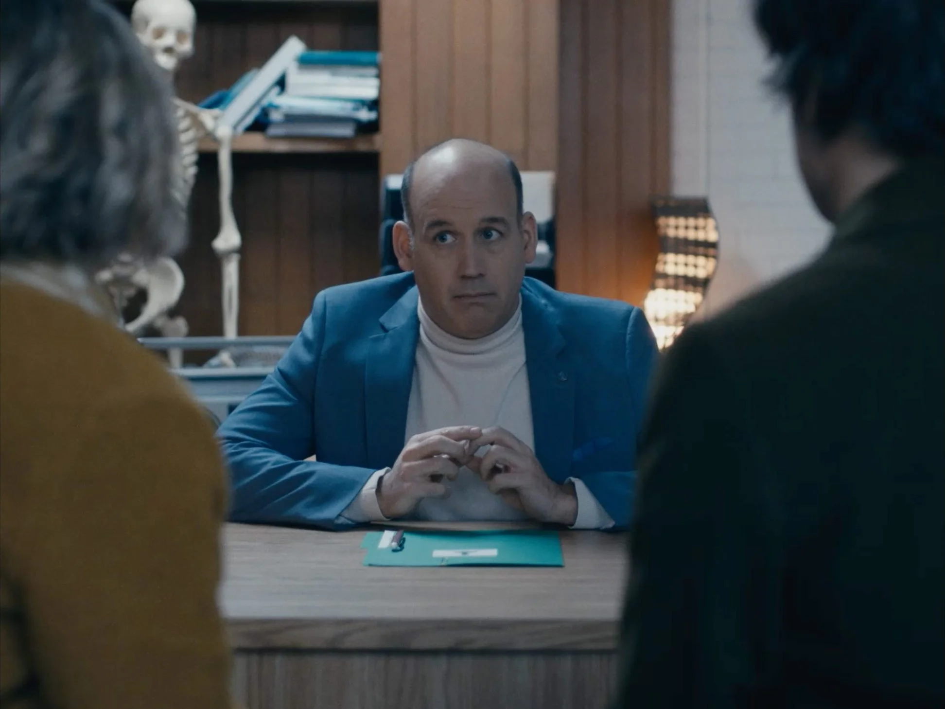 A man in a blue suit with a white turtleneck sitting at a desk, listening to two women on either side of him during an interview or meeting in a wood-paneled office.