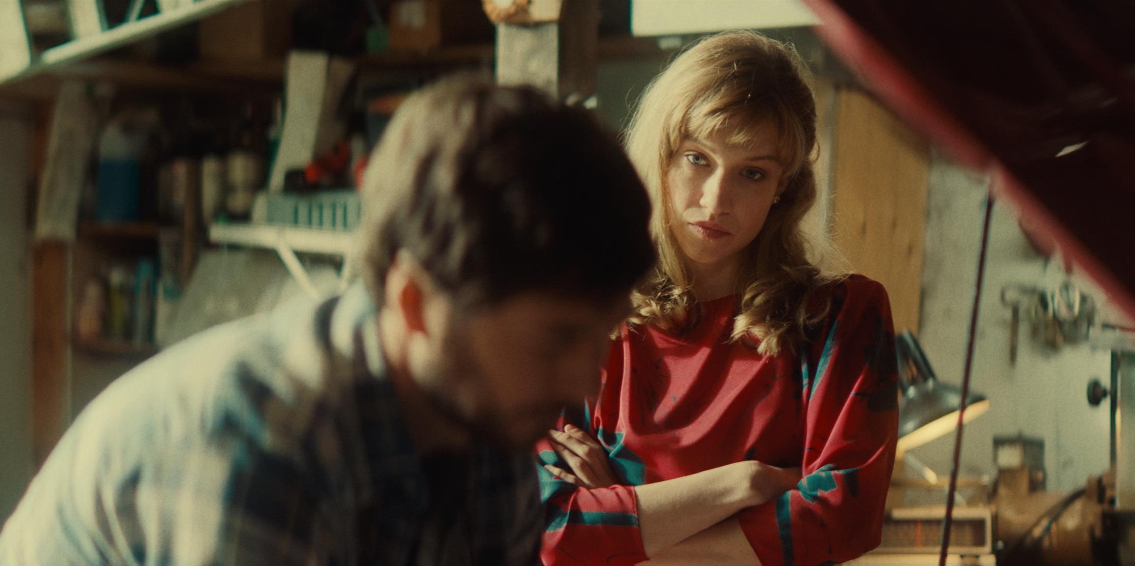 A woman with blond hair and a red shirt observing a young man with dark hair working on a project in a workshop or garage.