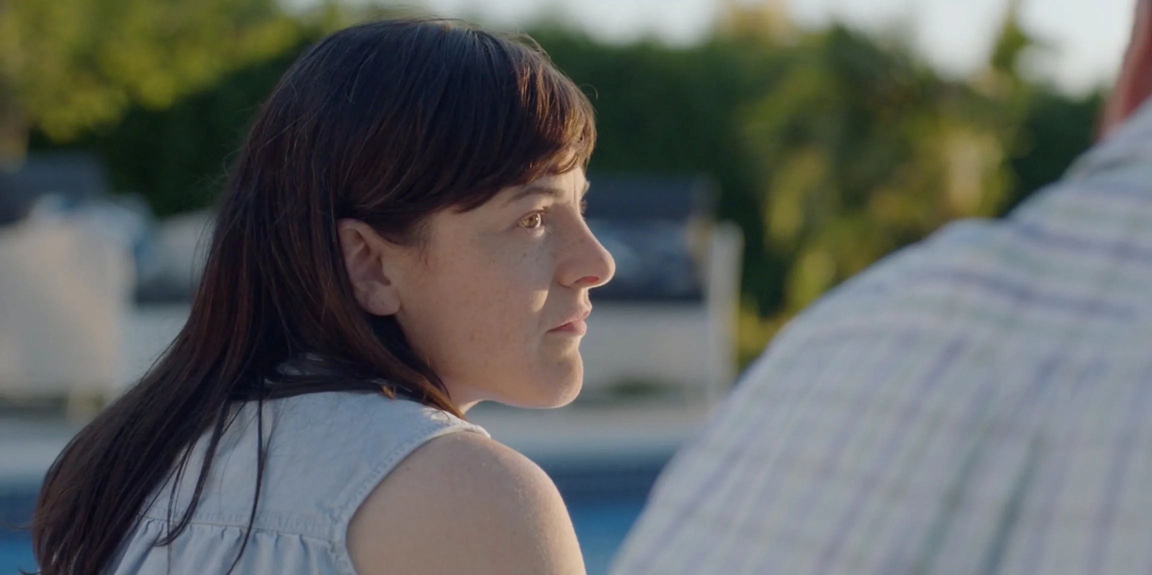 A woman with brown hair and fair skin looking thoughtfully at a man, outdoors during daytime with trees and a cloudy sky in the background.