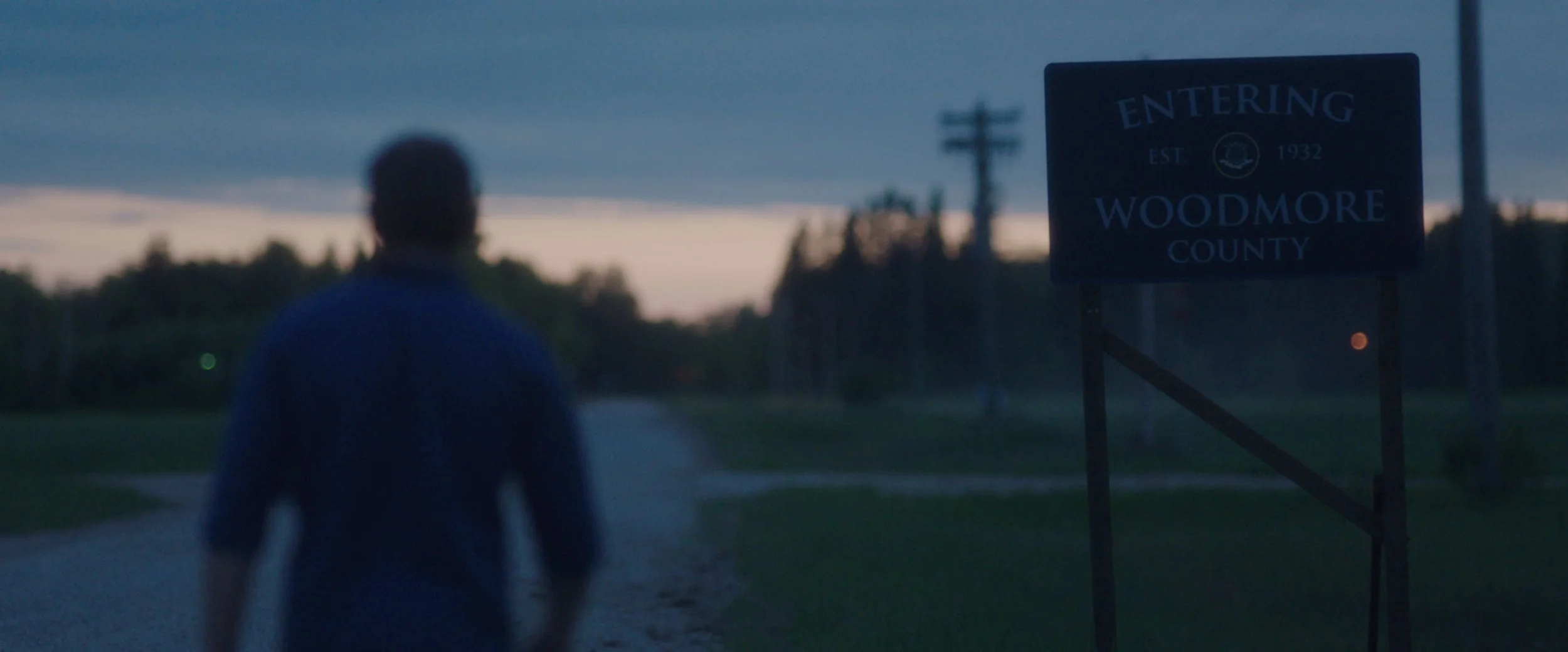 A person walking towards a sign that reads 'Entering Woodmore County, EST. 1932,' at dusk or dawn with a cloudy sky and trees in the background.