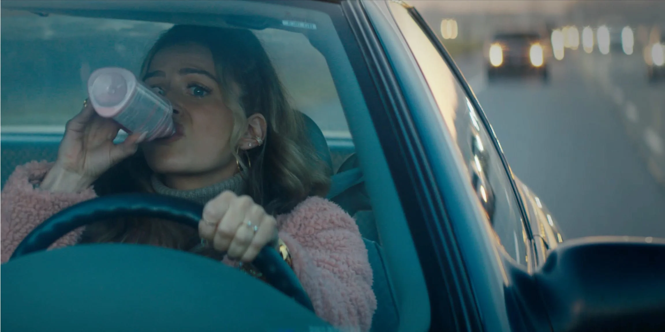 A woman with blonde hair wearing a pink fuzzy jacket drinks from a can while driving a car, with cars visible on the road outside.