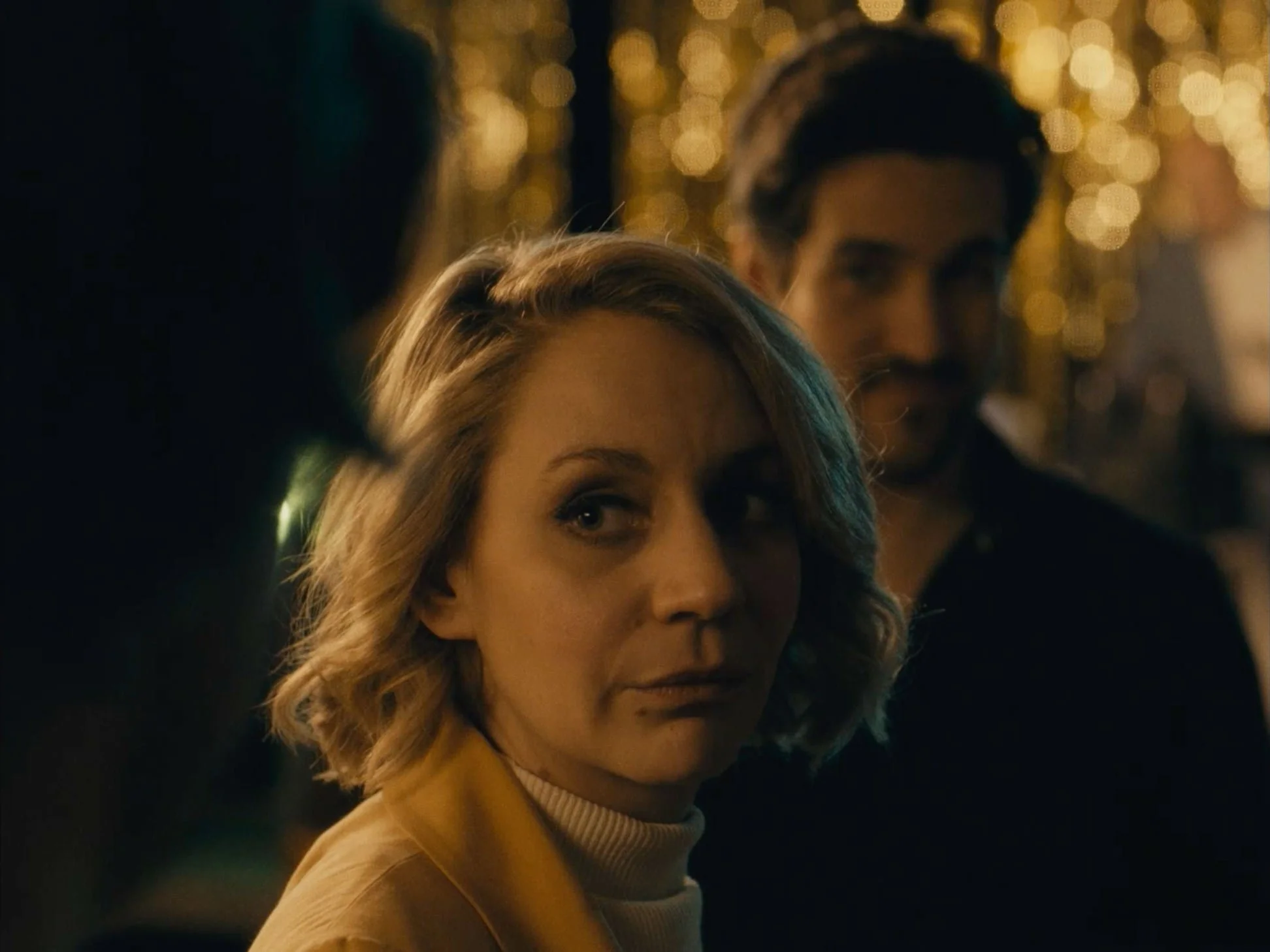 A woman with short blonde hair and a man with dark hair and a mustache, both looking sideways, in a dimly lit setting with warm bokeh lights in the background.