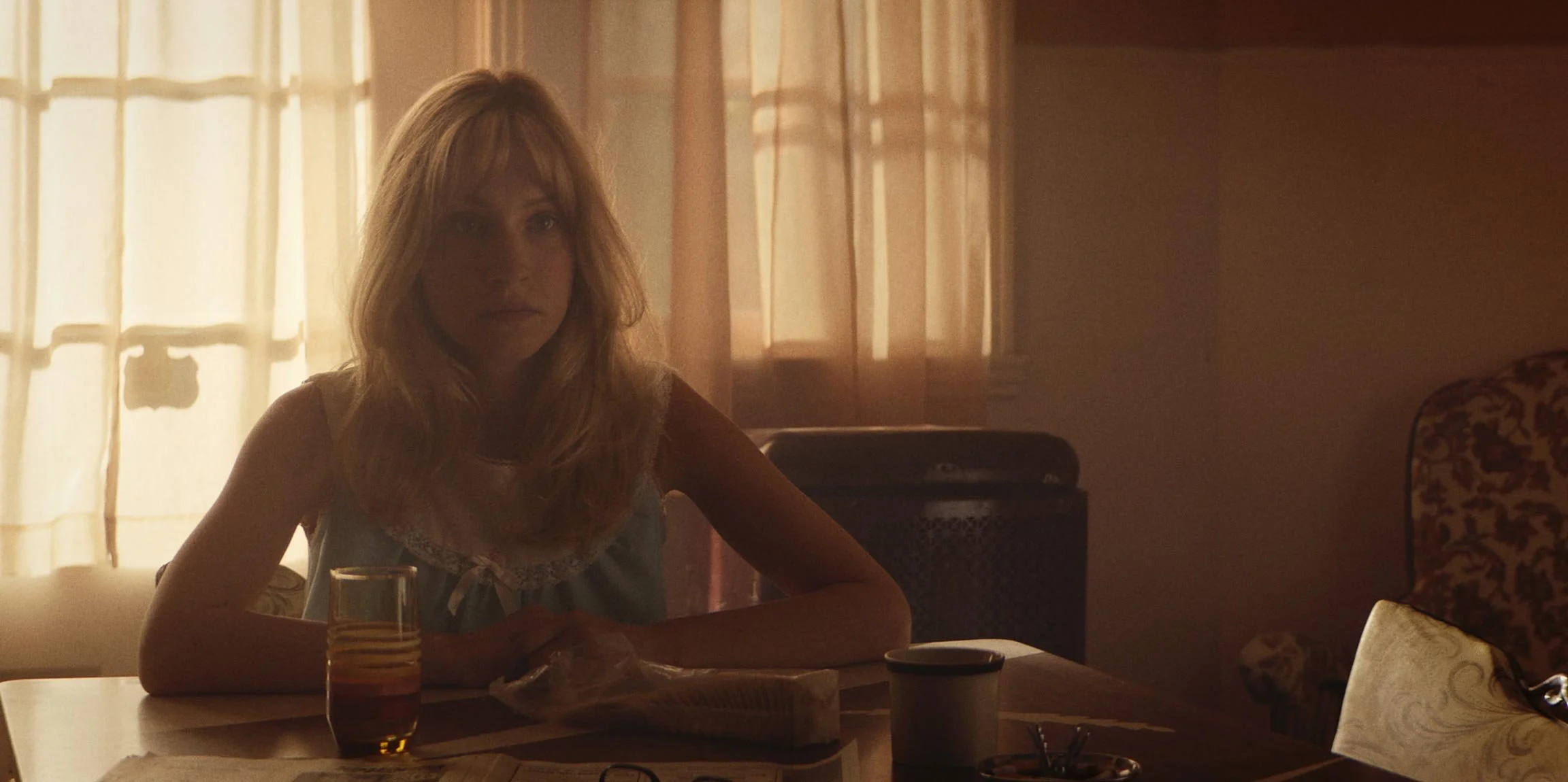 Young woman with blonde hair sitting at a dining table with a glass of tea and a cup, backlit by sunlight through curtains